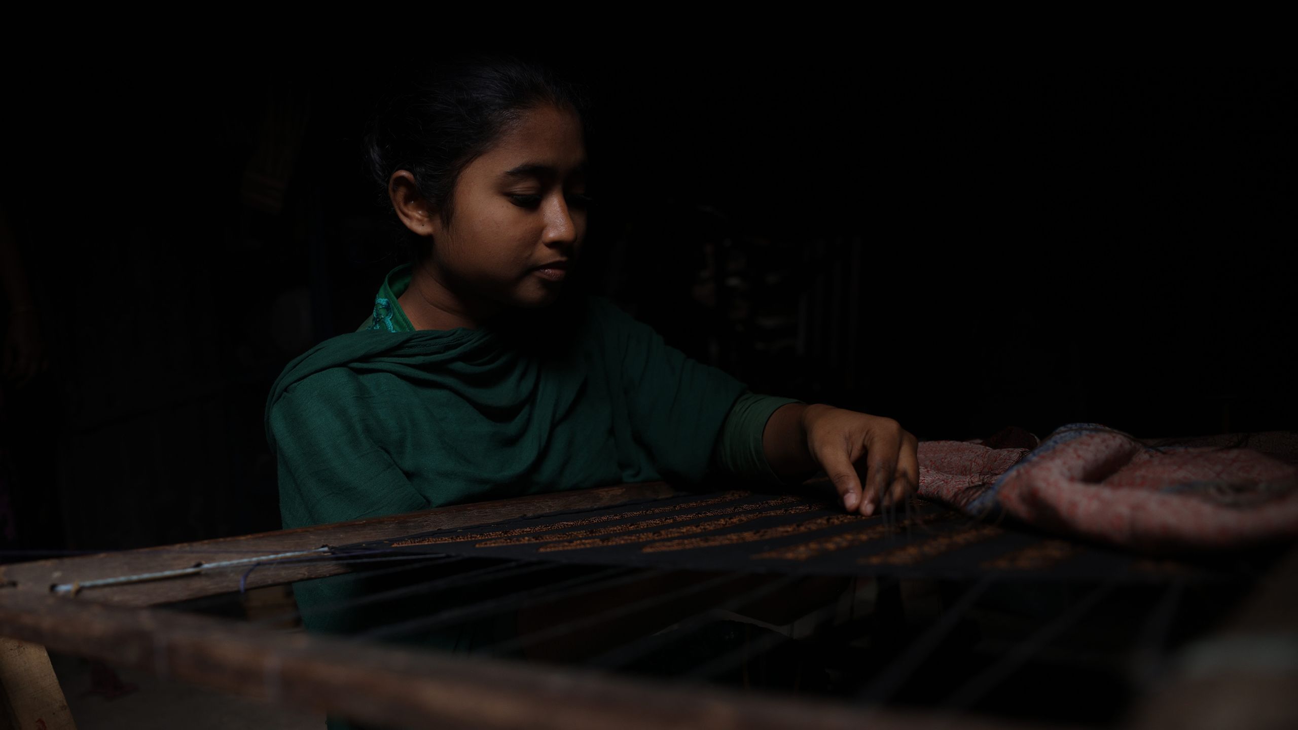 In a lightless room, a young woman in a green sari sits at a loom weaving cloth out of fine thread..