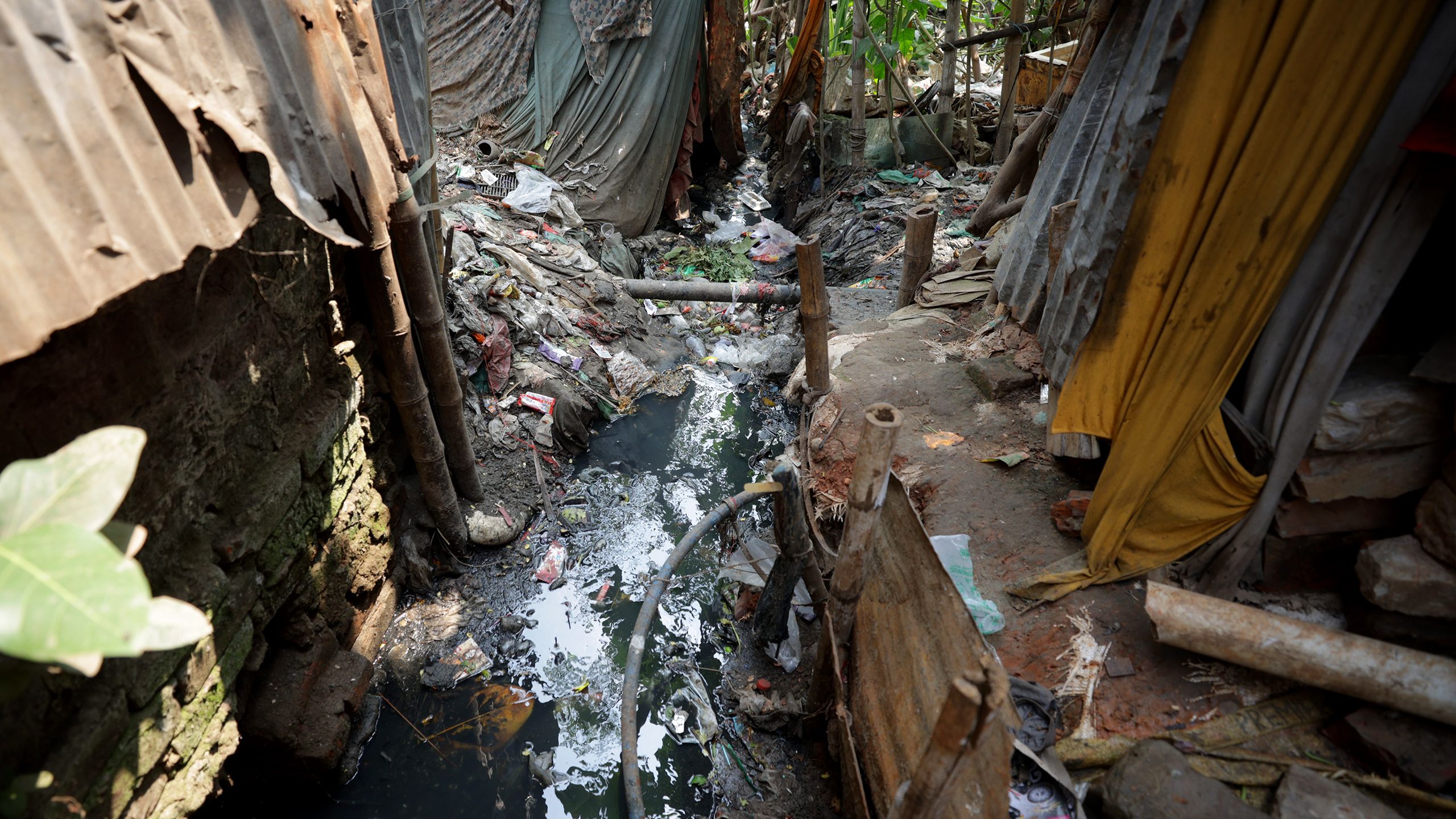 Standing water and sewage fill a gulley between homes, clogged with trash and debris. Water lines for the surrounding houses snake alongside the stream.