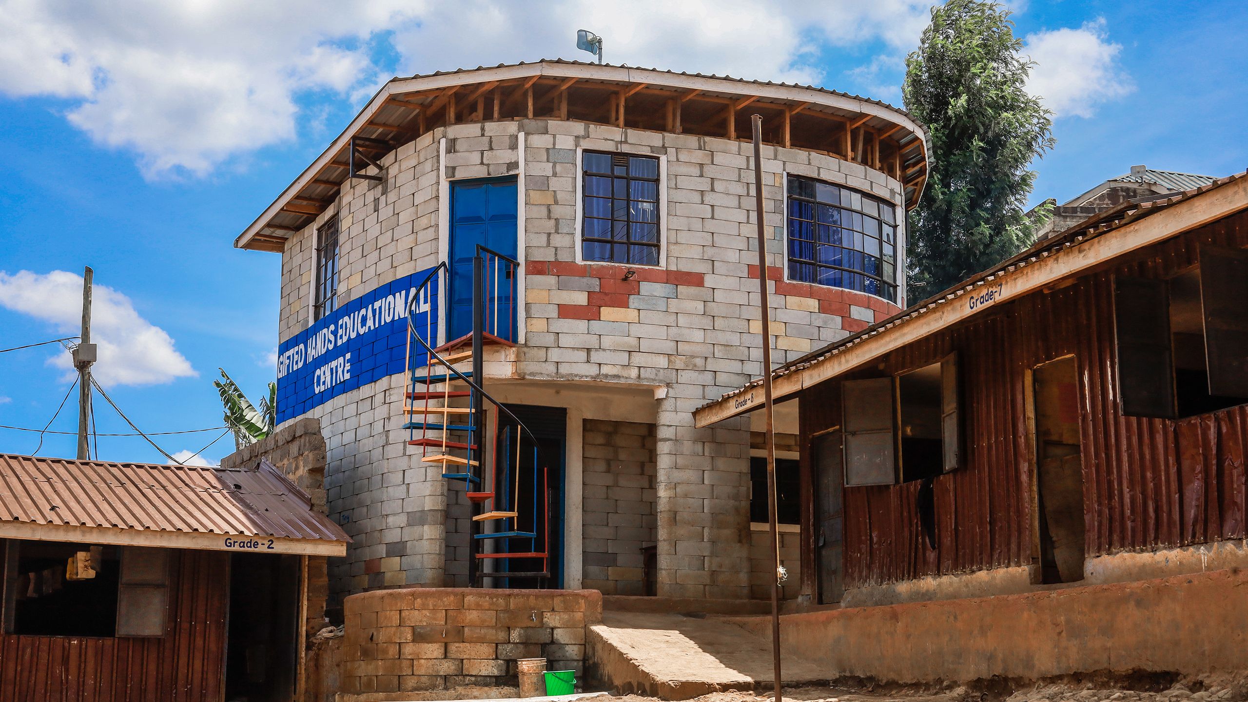 A school building built with Twistblocks. On the side is a painted sign reading "Gifted Hands Educational Centre." A multicolored spiral staircase leads up to the second story.