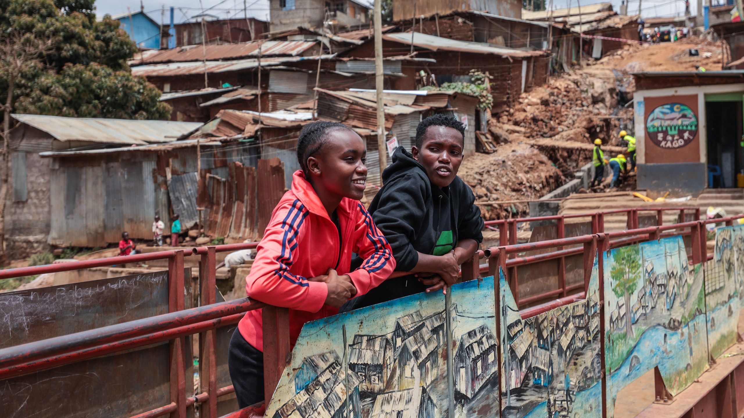 Milka and her daughter chat as they lean on the railing of a bridge over a ravine with a mural on the side. Behind them are houses built of rusting corrugated metal and crumbling bricks, as well as construction workers at work on a new building.