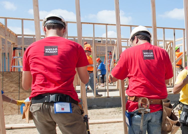 Two volunteers in hard hats and red Wells Fargo t-shirts, backs to the camera, help raise a framed wall on a Habitat build site.