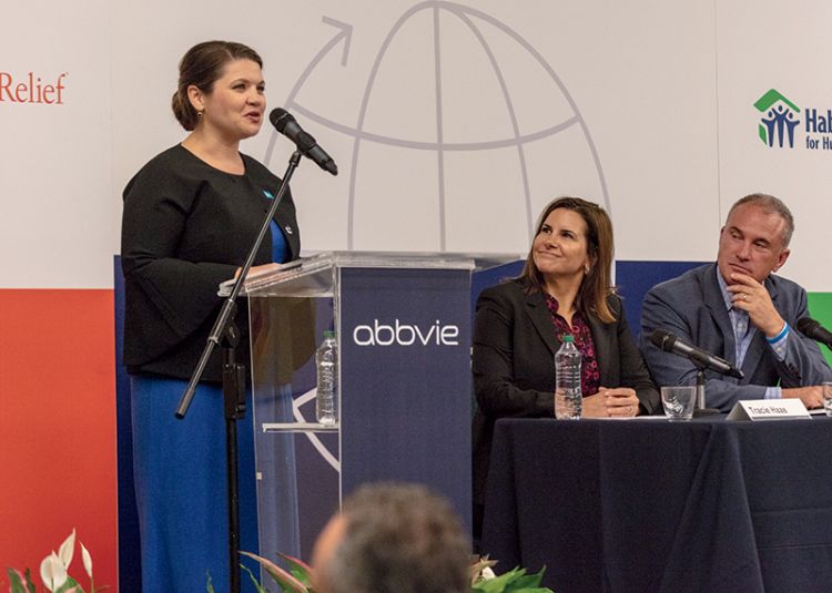 A woman in a black jacket and blue dress speaks from a lectern with the Abbvie logo on the front. To her left are a woman and a man seated at a table looking at her. Behind her is a wall with the Direct Relief and Habitat for Humanity logos.