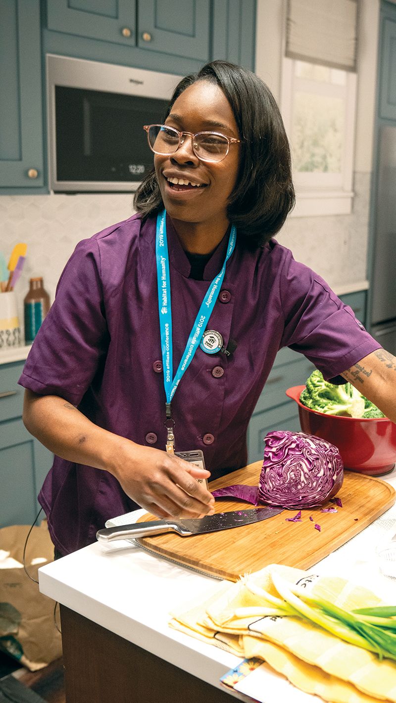 Chef Roshara stands at a counter with a cutting board and half-chopped cabbage, demonstrating a recipe