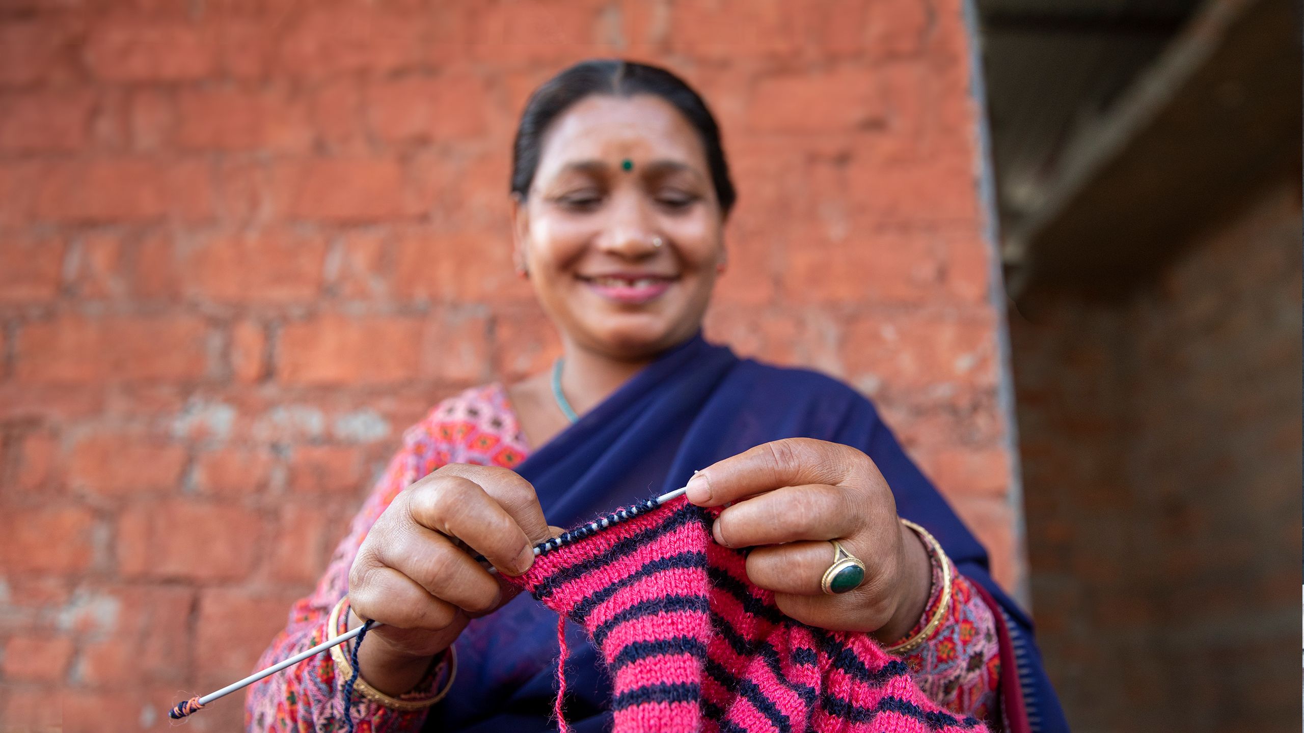 A woman in a sari knits a pink and navy striped sweater in front of a brick wall.