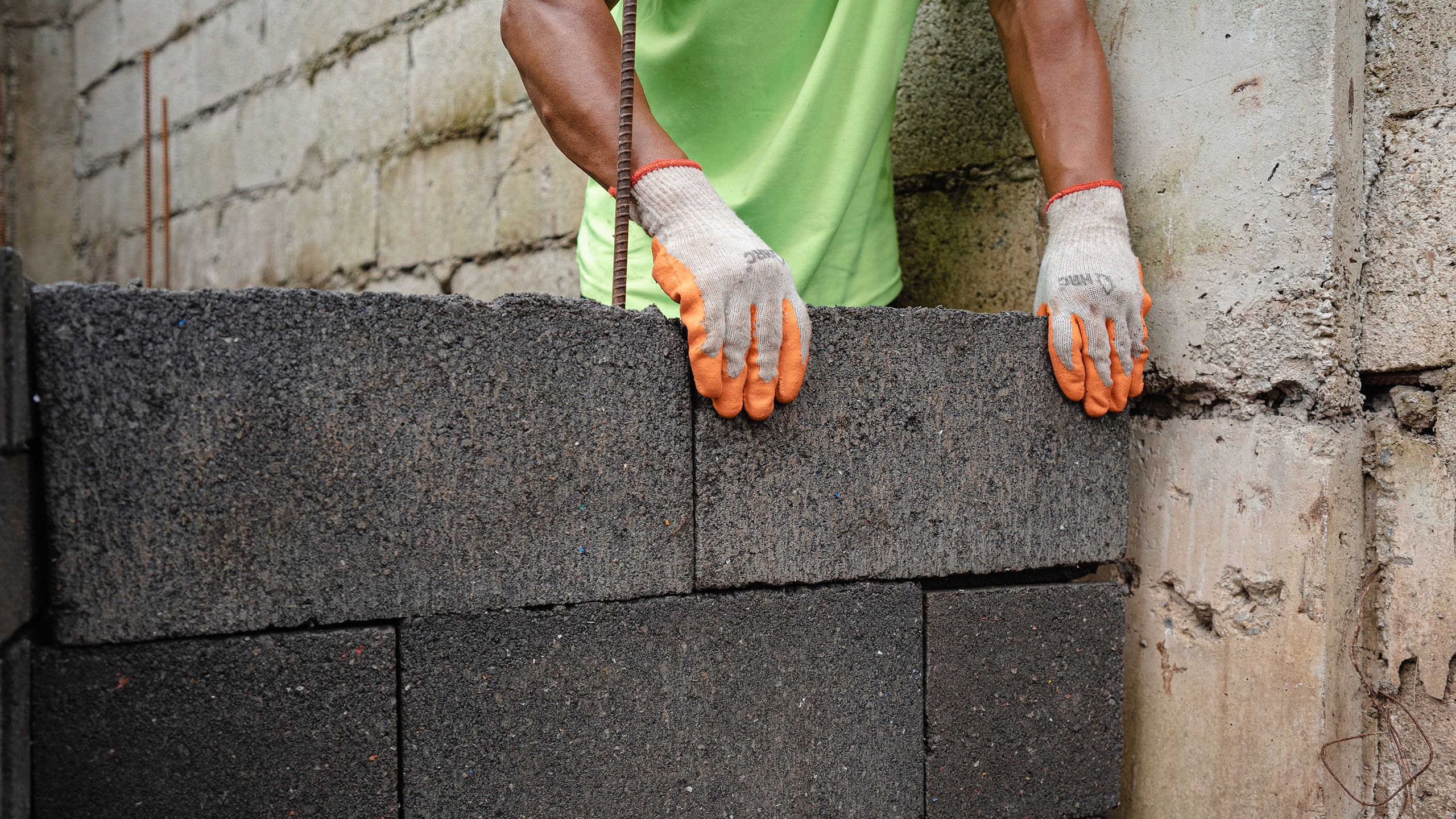 A person with bright orange gloves places a concrete block on a growing wall at the corner.