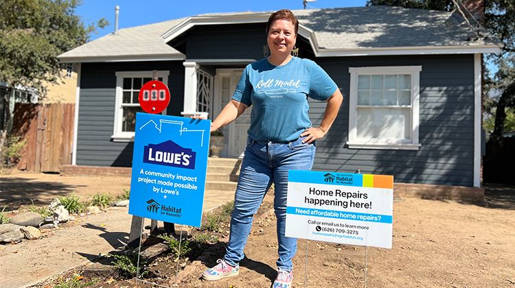Xochitl stands in front of her repaired home, her hand on a sign reading "A community impact project made possible by Lowe's."