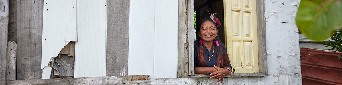 An older woman peers out from a window with pale yellow shutters, smiling.