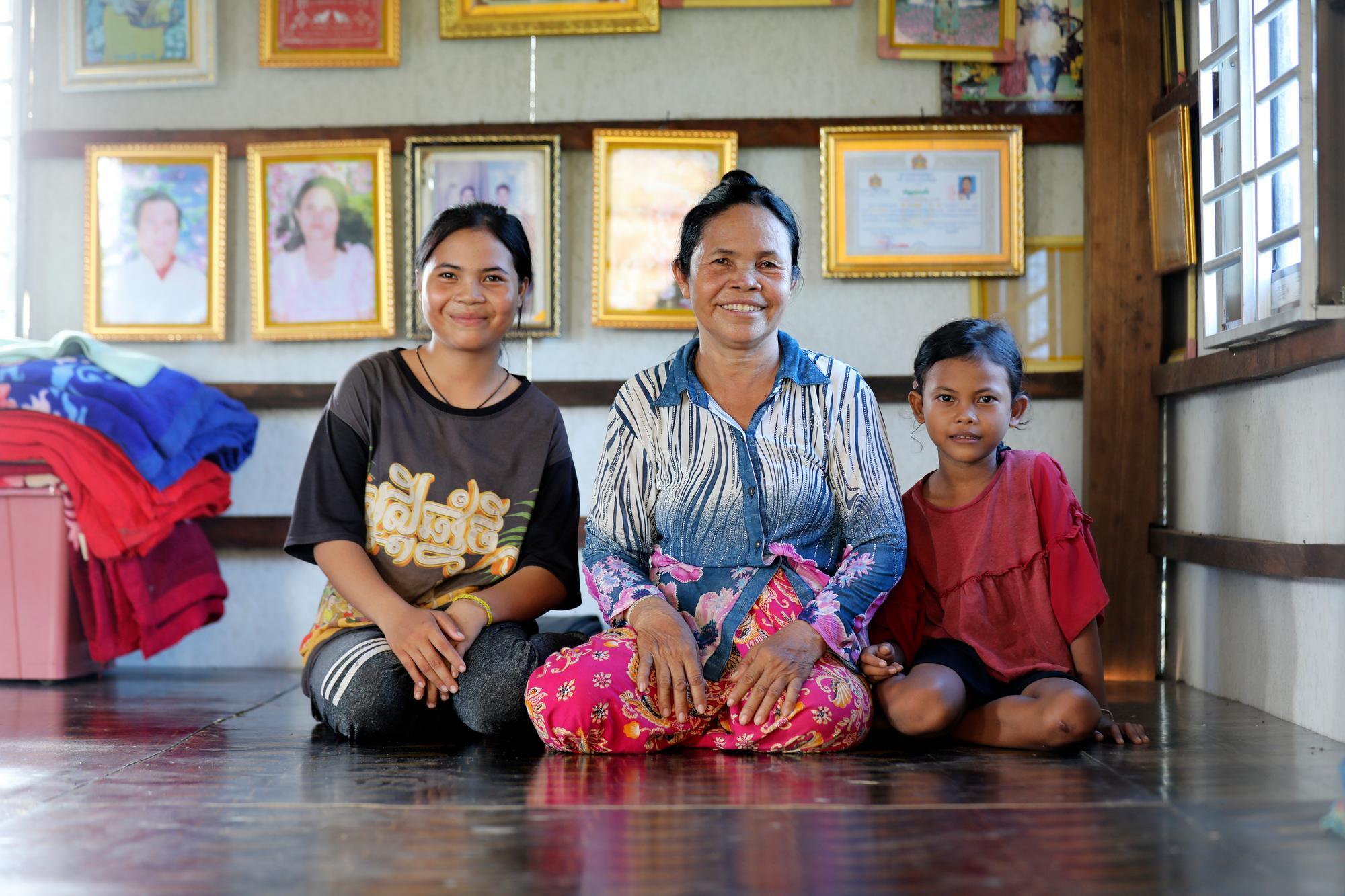 Rem is flanked by two of her granddaughters, seated on the floor in their home in Battambang, Cambodia. Behind them a wall of gold frames displays family photos and awards.