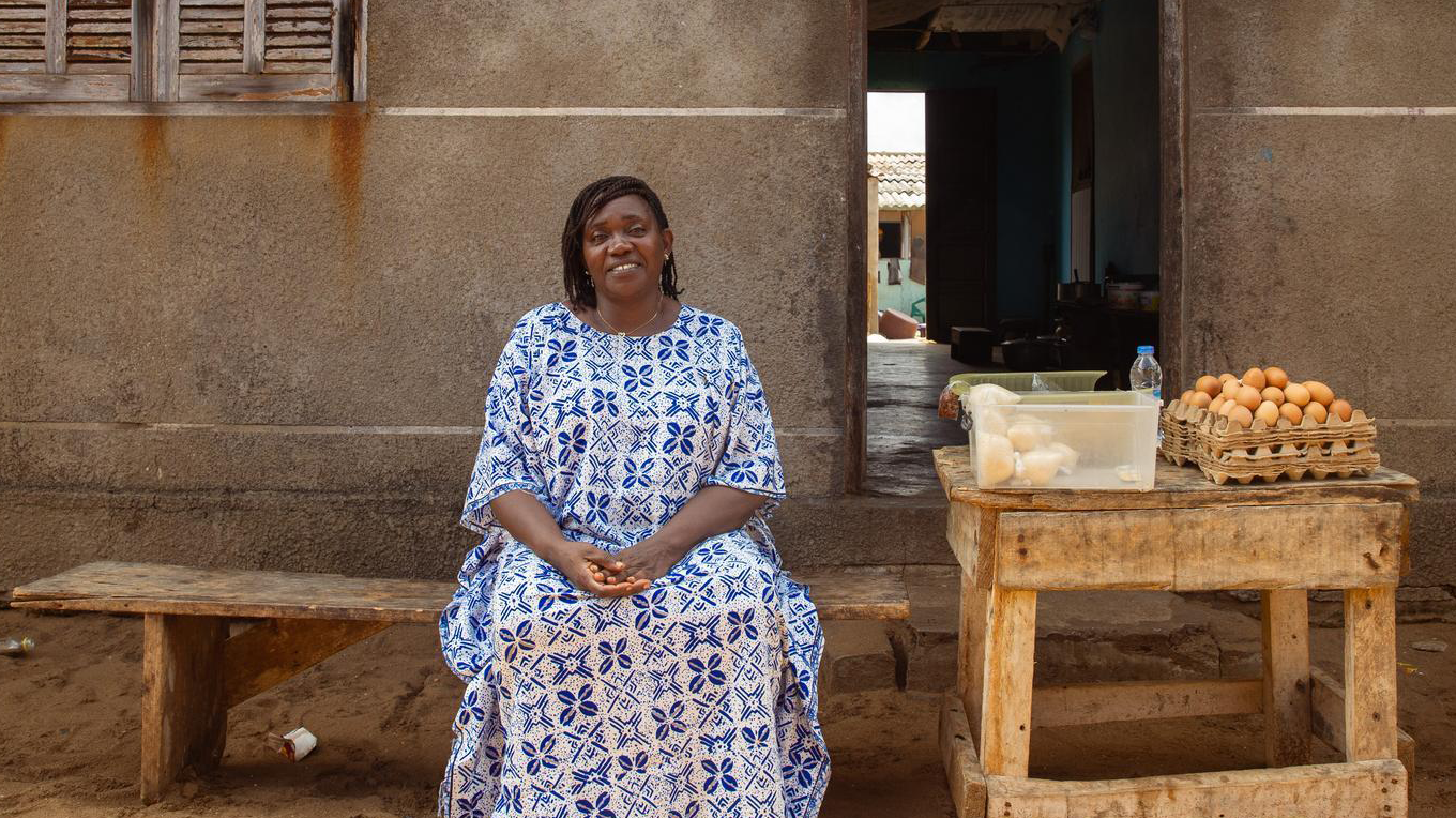 A woman in blue sits on a bench outside her home with her hands clasped in her lap.