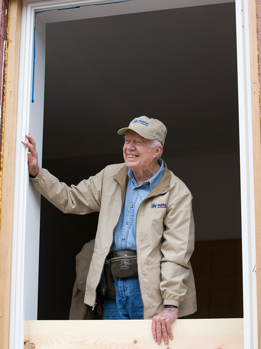 President Carter smiling at someone off camera, standing framed by a window on a build site.