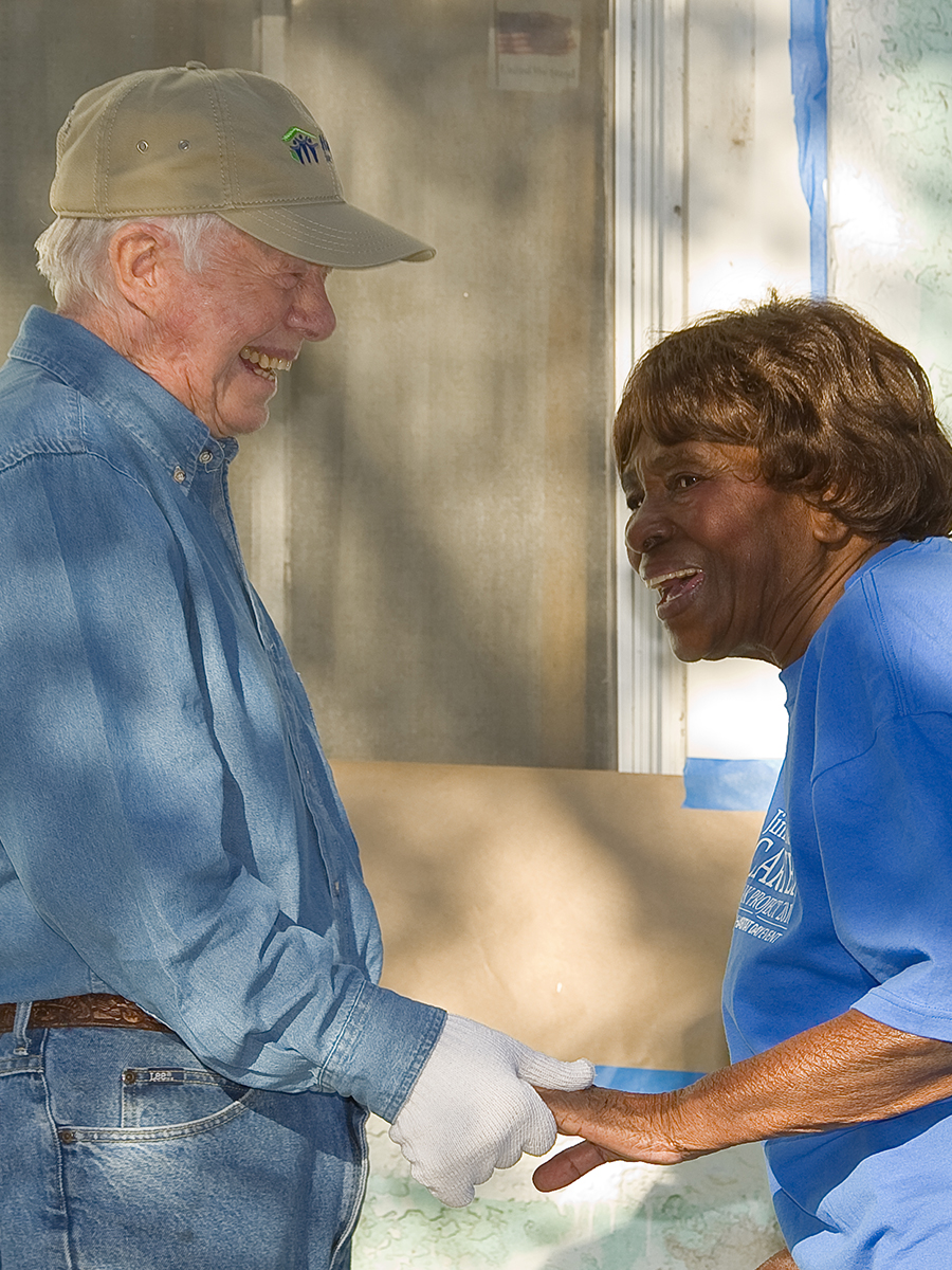 President Carter holding the hands of a homeowner as they talk and laugh, taking a moment during a build.