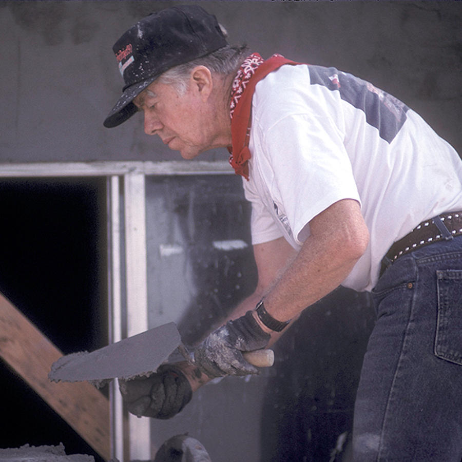President Carter in a hat and bandana, bending over to work on helping pave the foundation.
