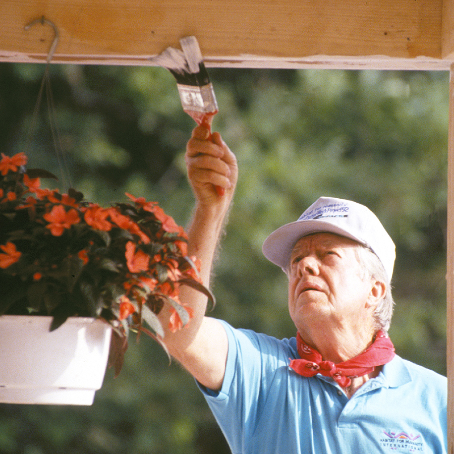 President Carter painting a porch roof, wearing a cap and red bandana.