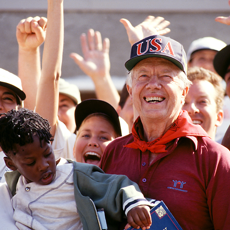 A photo of President Carter holding a child, smiling in a crowd who's raising their arms in celebration.