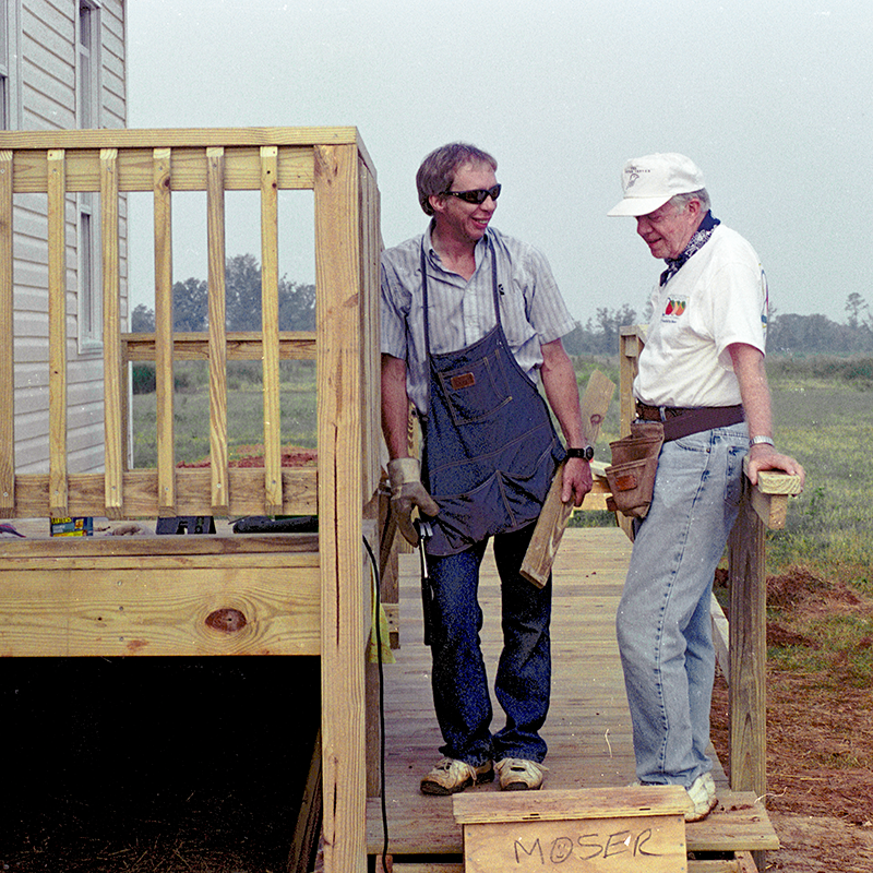 President Carter on the porch o a build site smiling with a volunteer, taking a moment from building to talk.