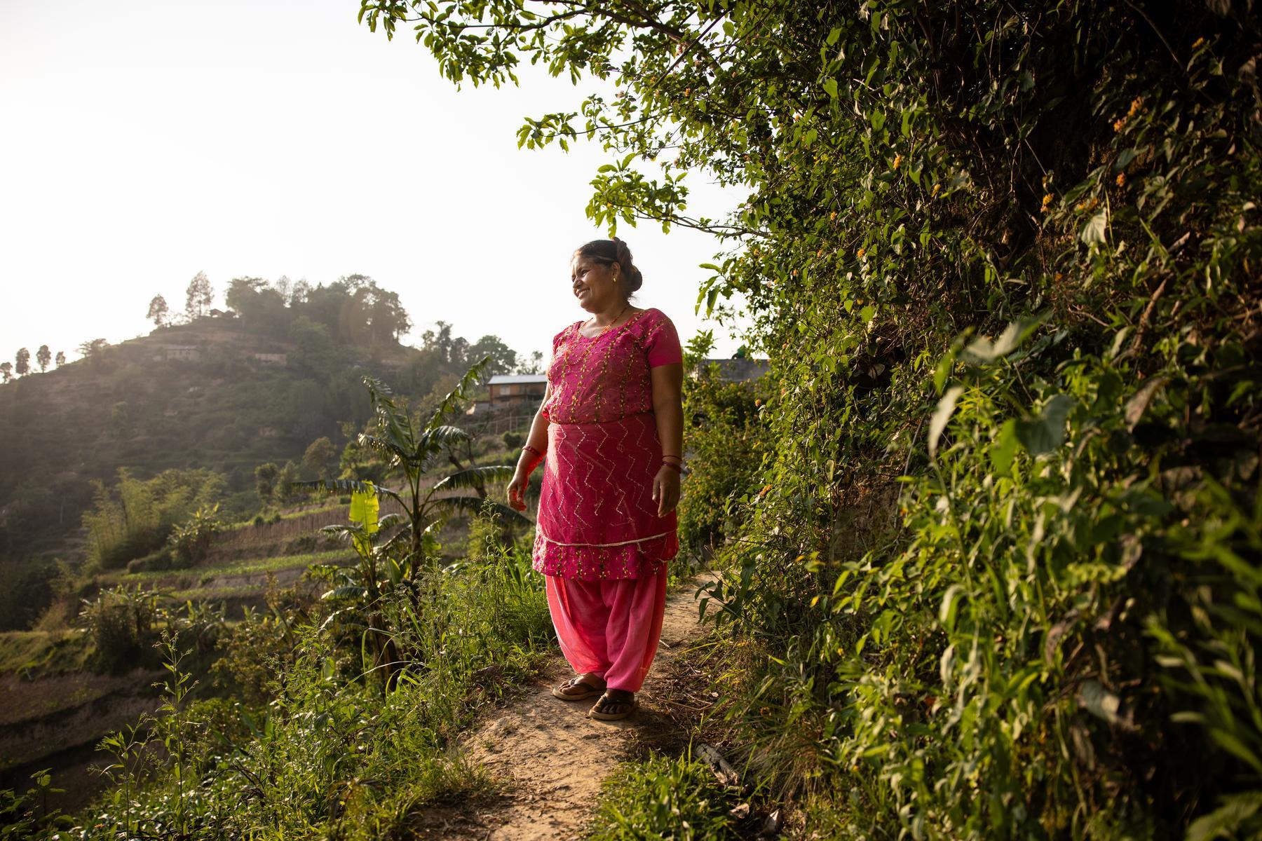Niru walks a path near her house. She is surrounded by green trees. She is wearing a pink sari and looking off into the distance.