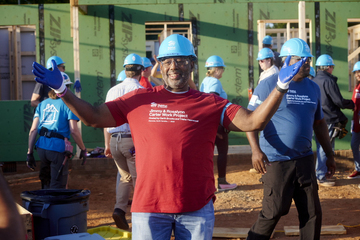 Smiling volunteer with arms outstretched in front of in-progress house