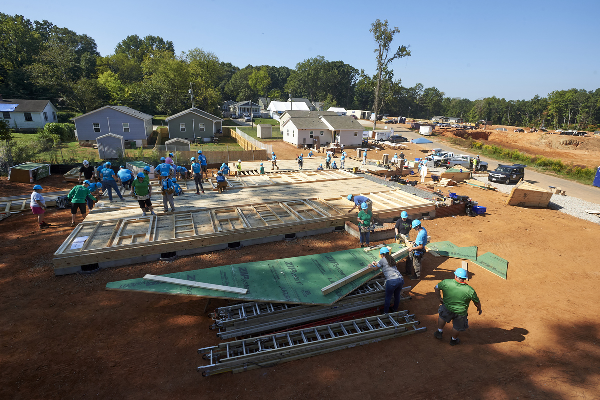 Wide view of construction site with some volunteers standing on completed wooden base while some work on assembling another part of the home.