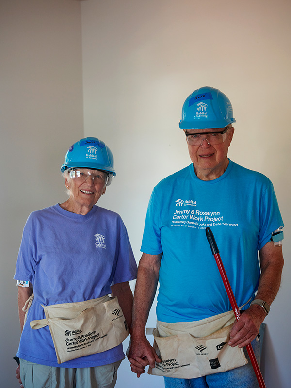 Married couple Jean and Gene stand in a white painted room, holding hands.