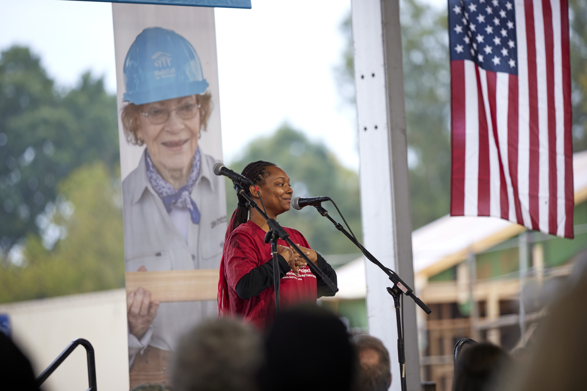 Future homeowner speaking to CWP volunteers in front of American flag and poster of Mrs. Carter.