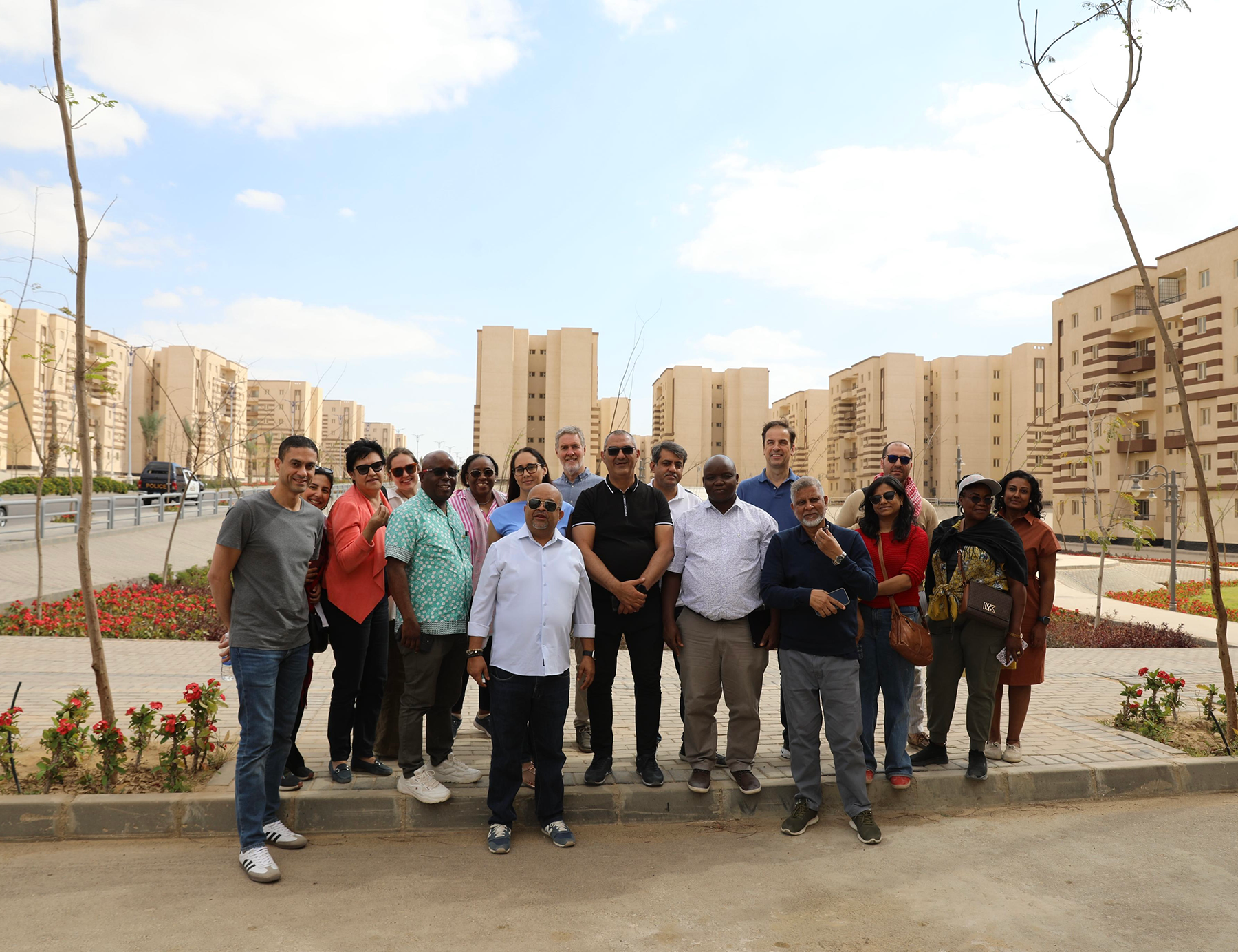 A group of people stand on sand-colored pavers and smile for the camera. A set of buildings are on display behind them.