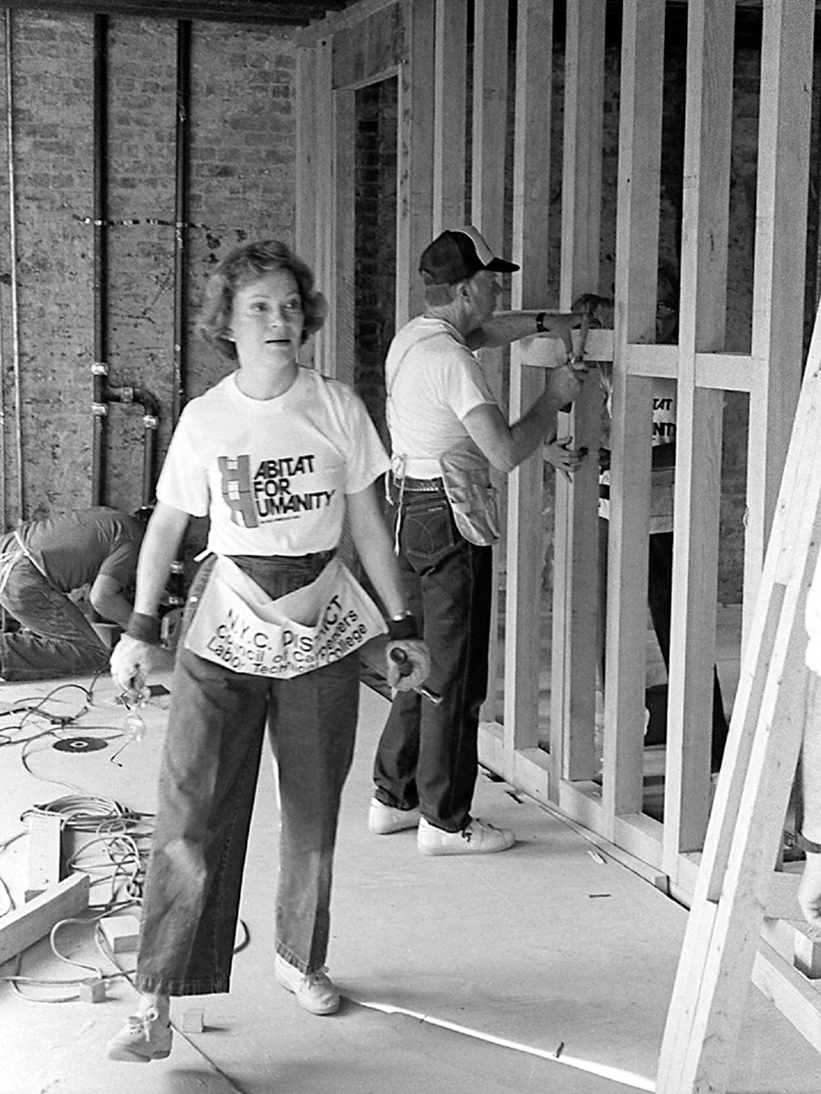 Black and white photo of the Carters inside a home under construction, Rosalynn is in the foreground and President Carter is hammering in the background.