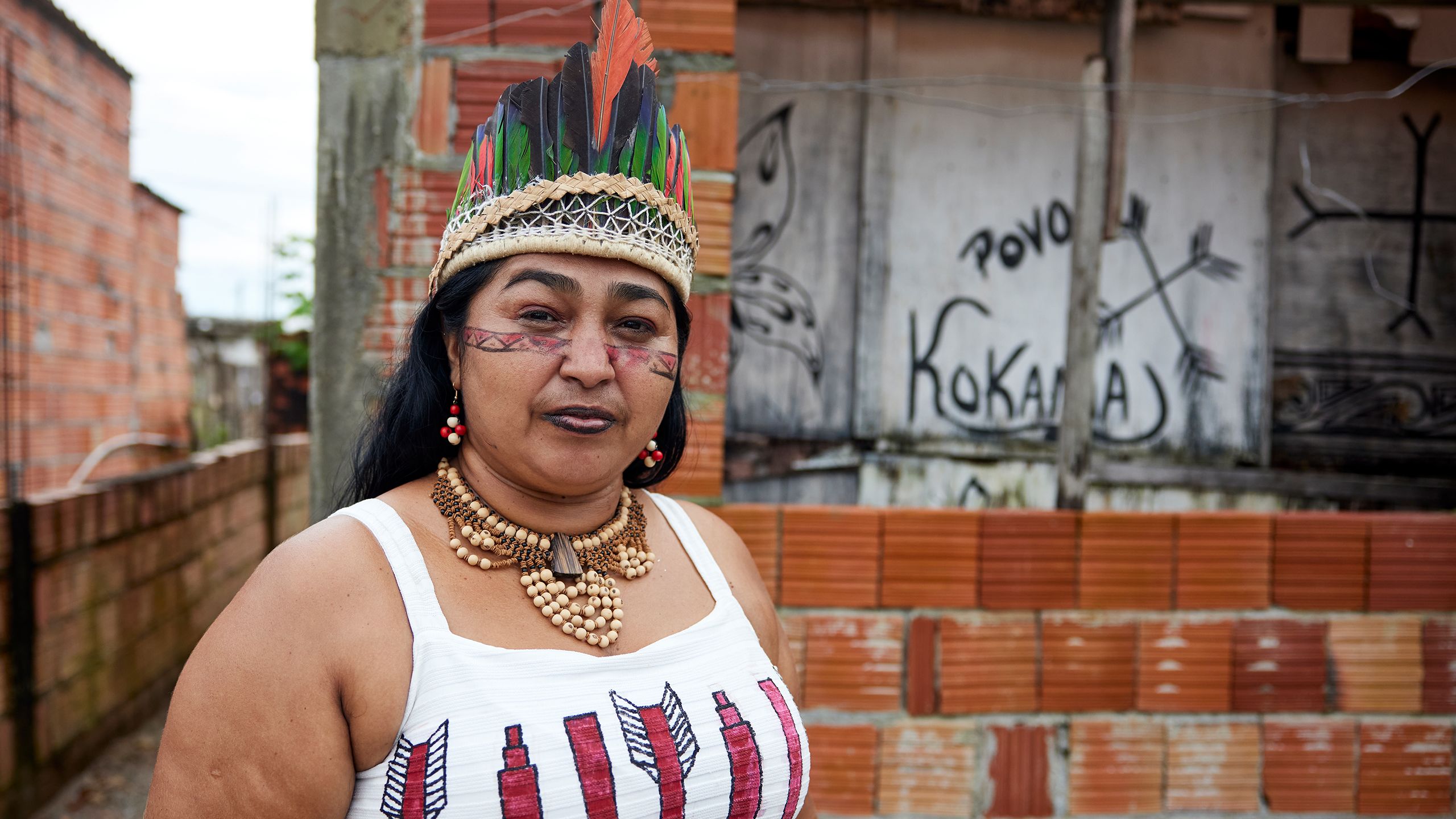 Sol, a young indigenous Brazilian woman, wears a traditional parrot feather crown and face paint. Behind her is a brick building with the word "Kokama" and a butterfly painted on a plywood-covered window.