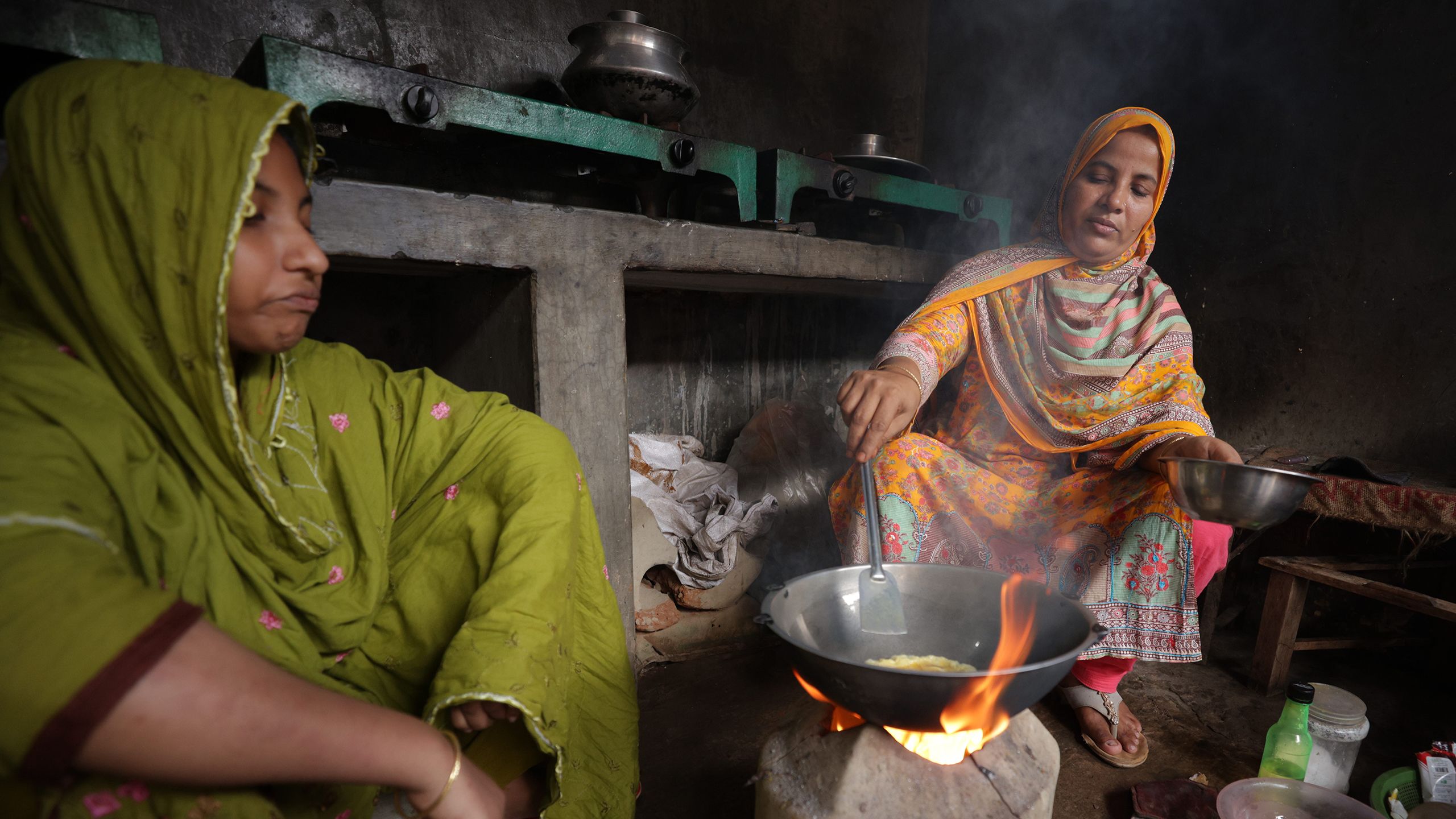 Sahana, a middle-aged woman in a sari, cooks a meal on a small ceramic stove in an indoor cooking facility. A neighbor sits with her, and smoke rises from the pan to cover her face.