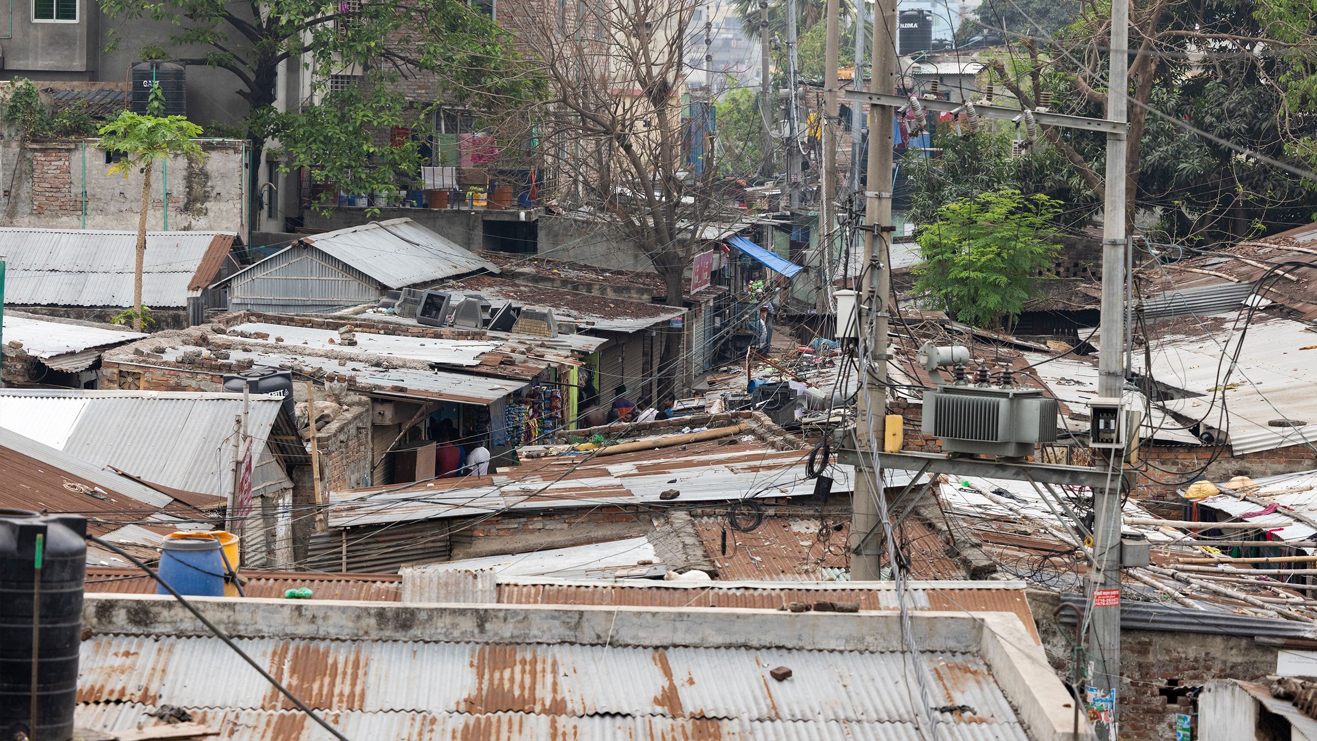 Roof-level view of houses and buildings with rusting corrugated metal roofs and a narrow pathway with tangled overhanging power lines.