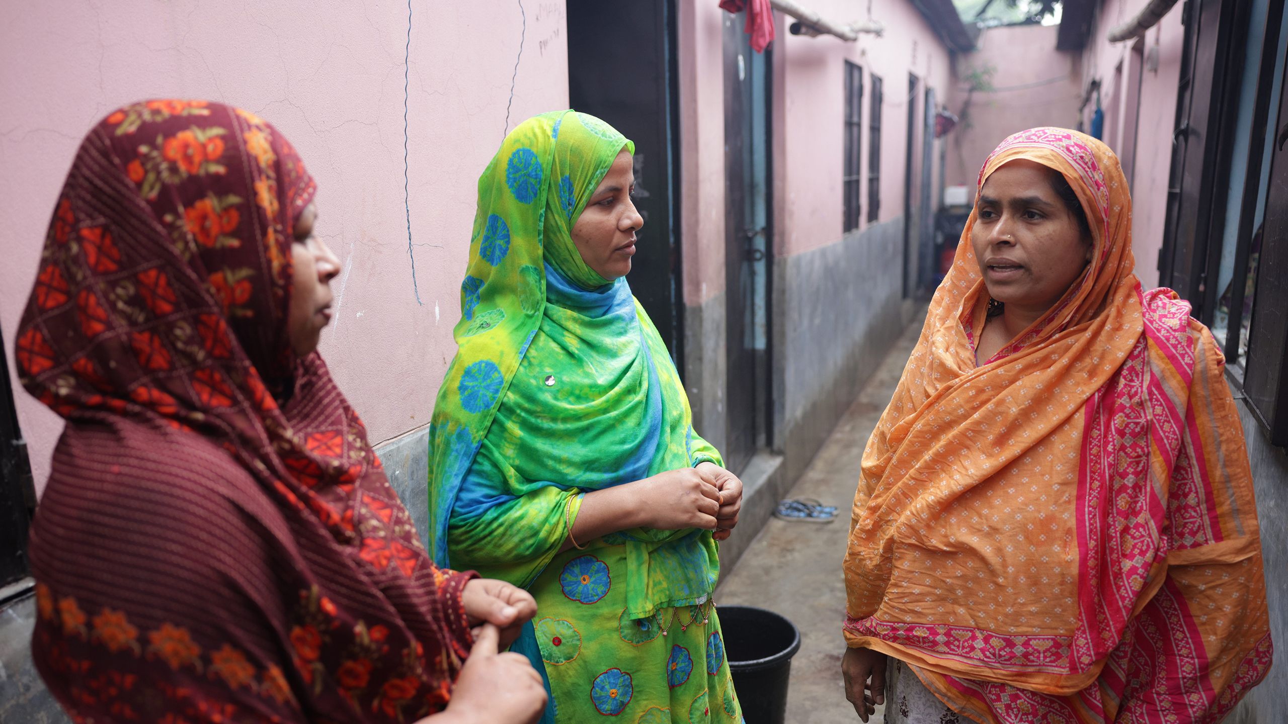 Sahana and Sumi converse with a neighbor in a pathway between their homes. the neighbor speaks as Sahana and Sumi listen intently.