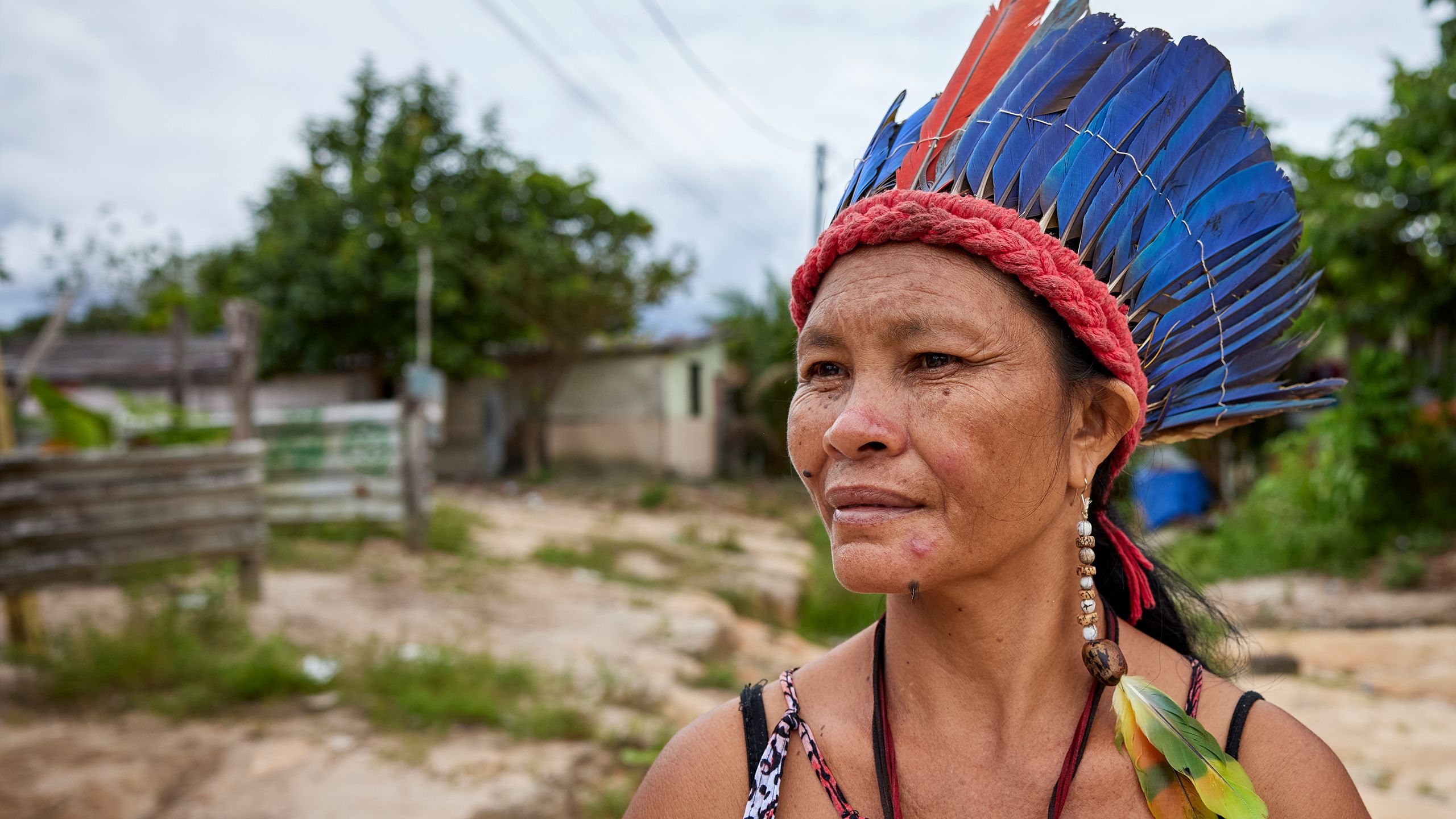 Lutana, an older Kokama woman, stands in a clearing near some homes. She wears a traditional macaw feather crown and green feather earrings.