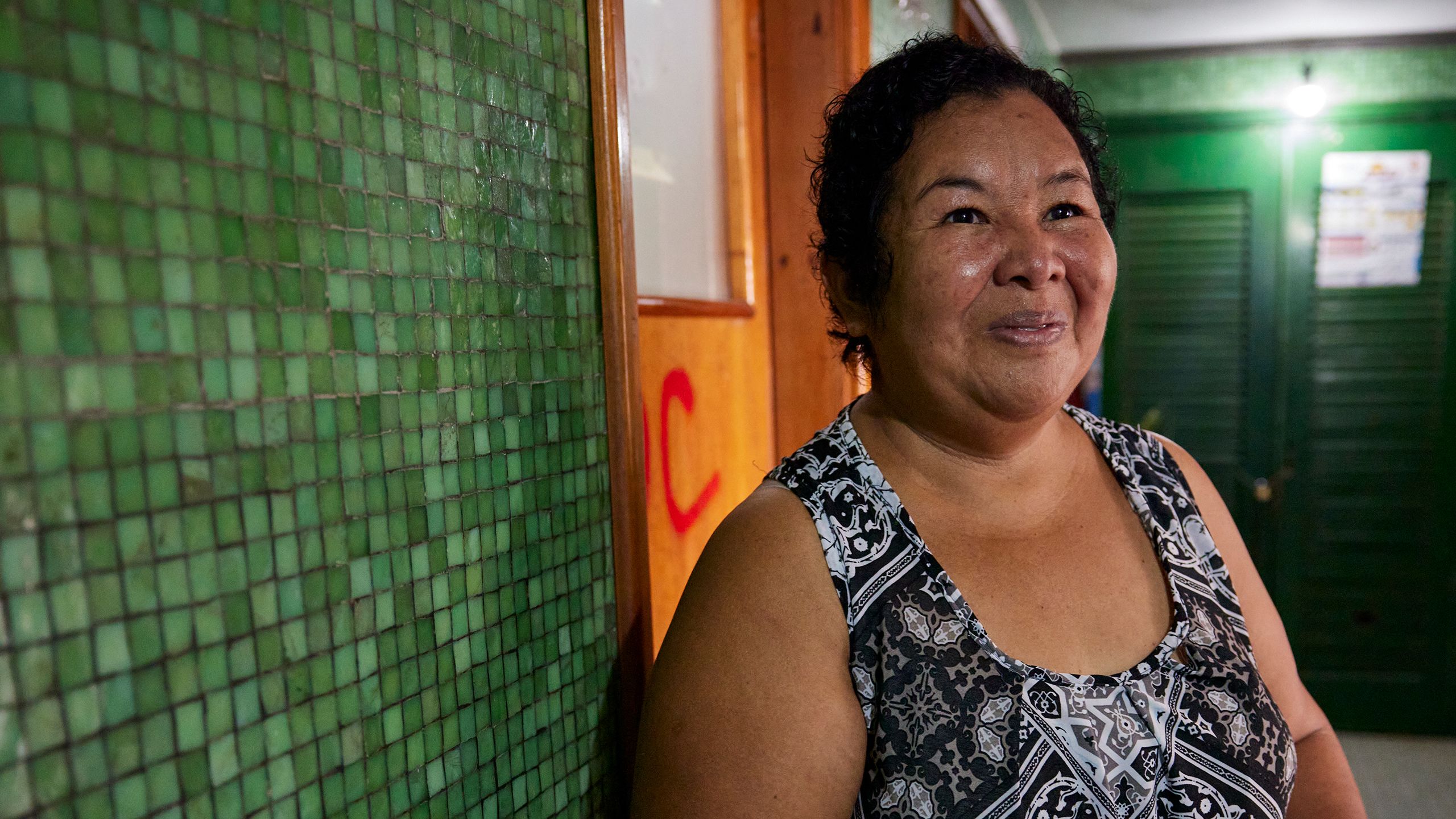 Vilma, a middle-aged Brazilian woman with short hair, stands in the green-tiled hallway of her apartment building