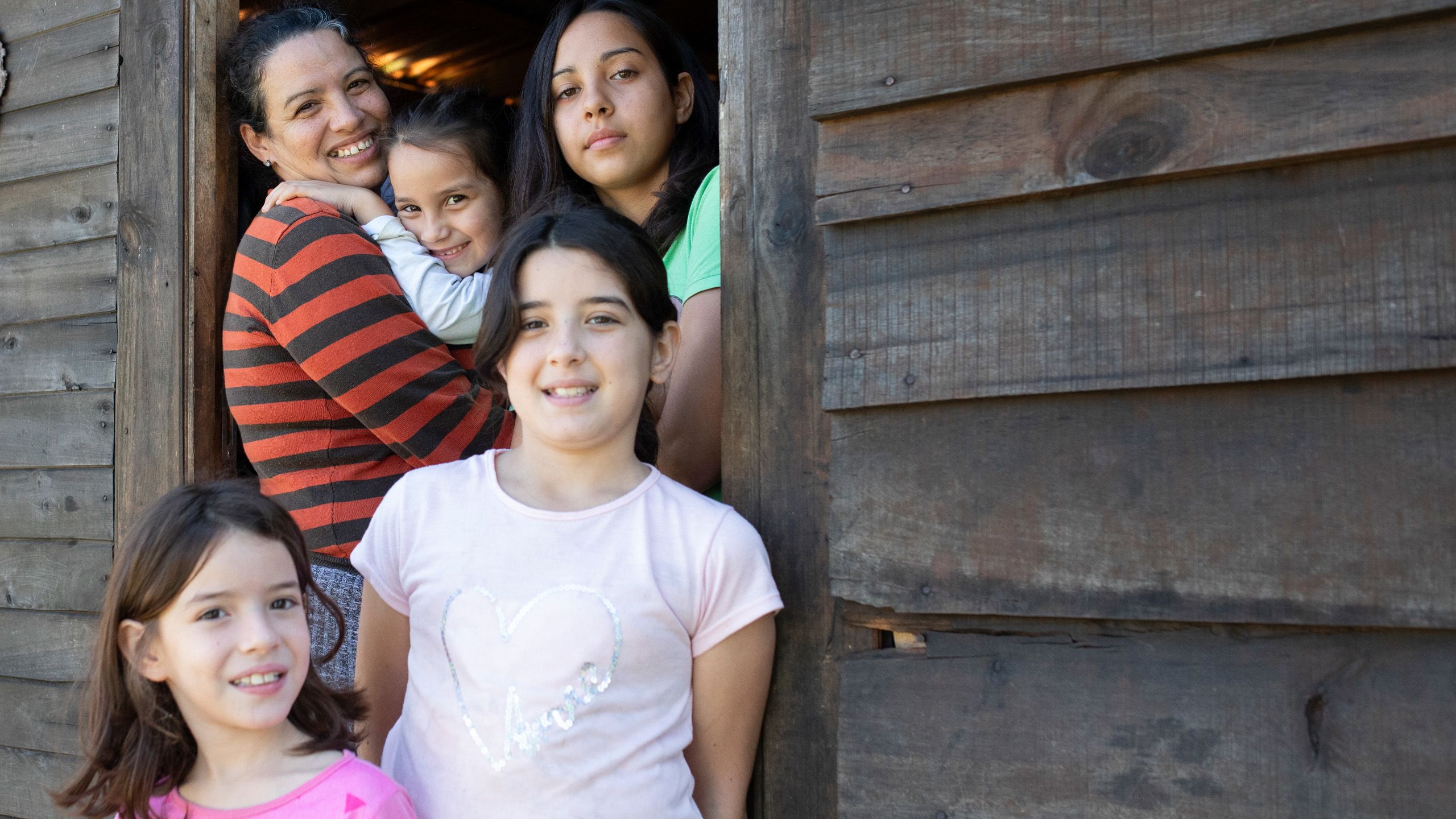 Alejandra holding her youngest daughter in a hug and smiling surrounded by her other three daughters in the doorway of her home.
