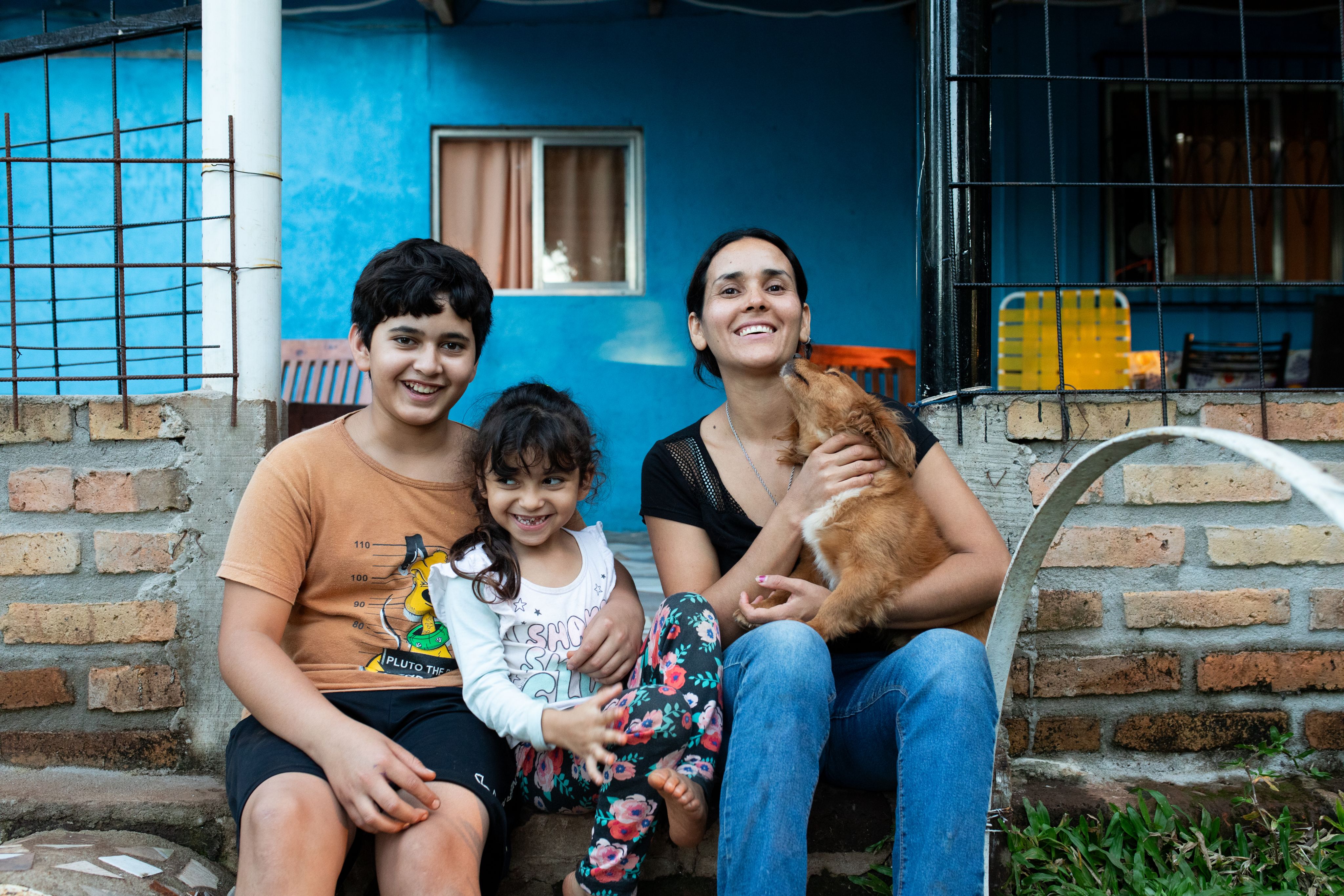 Portrait of Patricia, her son and daughter, and one of their dogs in front of their blue house.