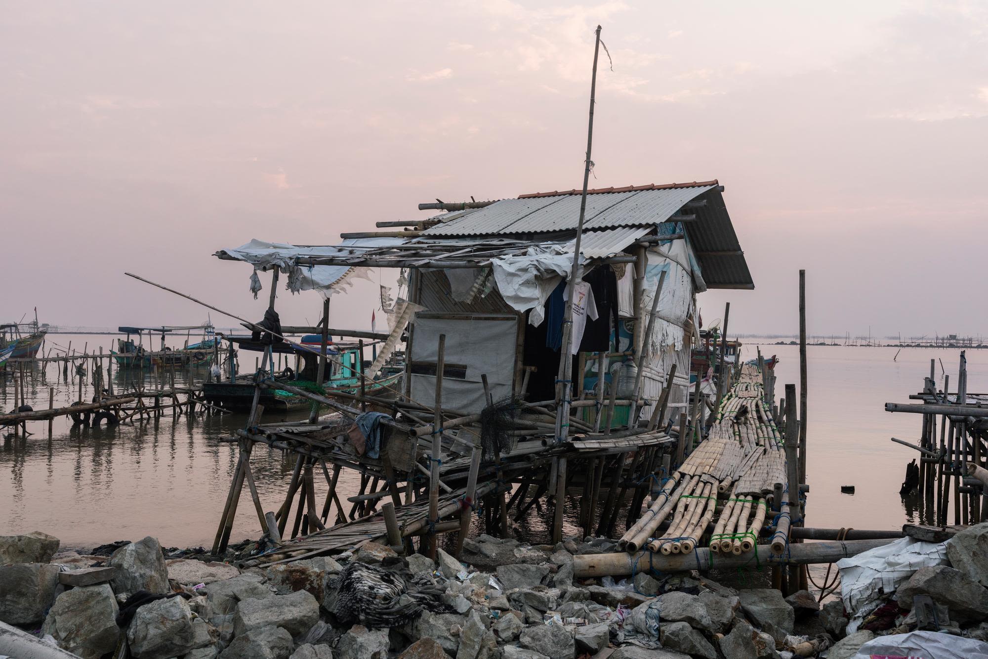 A rough, improvised shelter by the water, with fishing boats and a dock in the background.