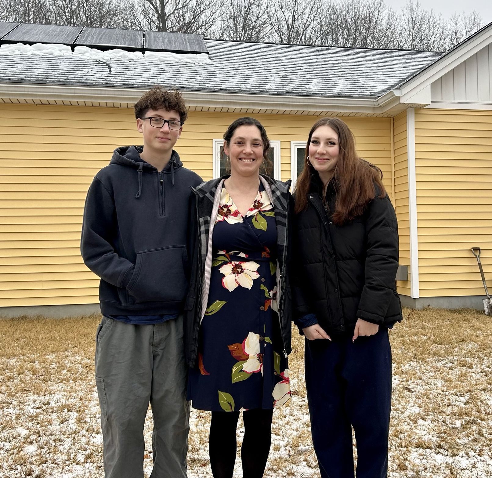 Aliyya standing with her two children in front of a yellow house with a bright blue door and solar panels on the roof.