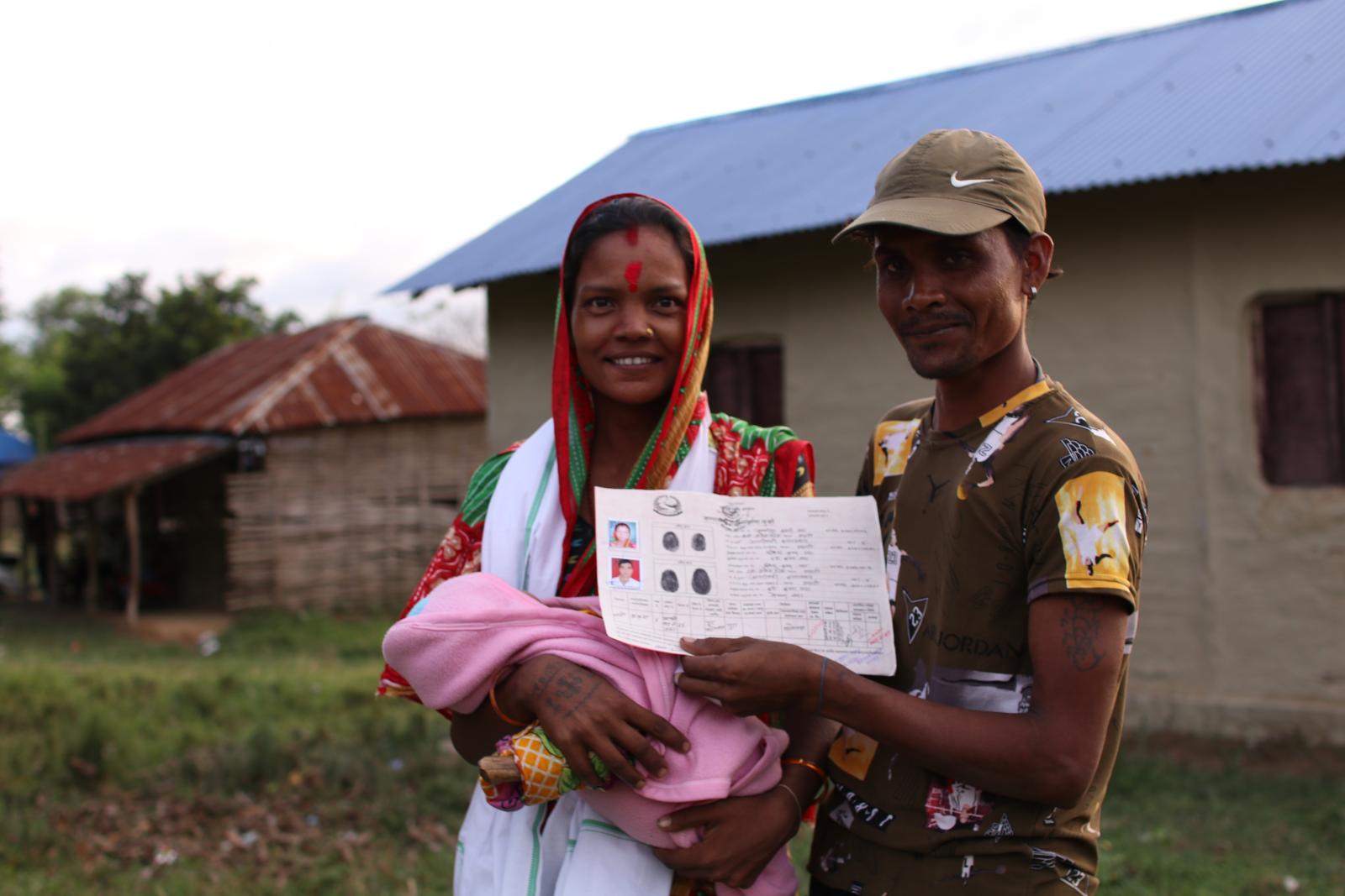 Smiling parents with a baby in front of their home. The man holds a land tenure document. 
