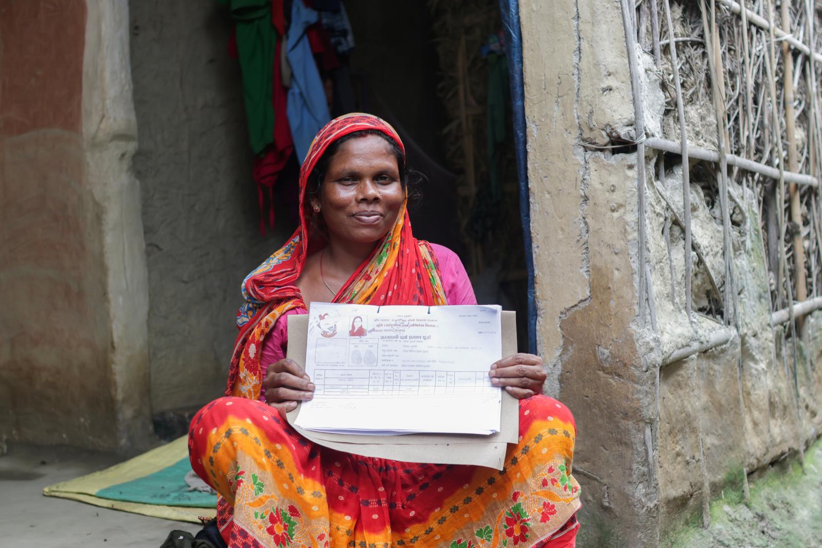 Woman in red, pink and orange garment sitting cross-legged on her front stoop. She is smiling and holding land tenure documents.