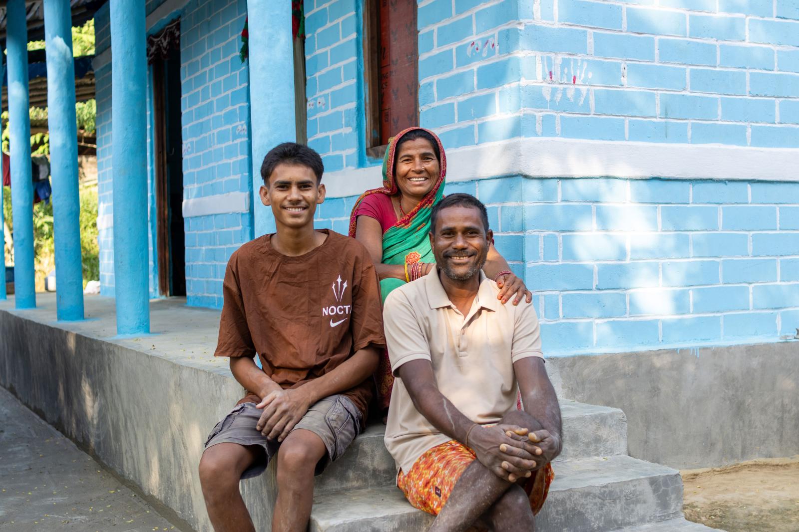 Smiling family of three sitting in front of their light blue painted brick house. 