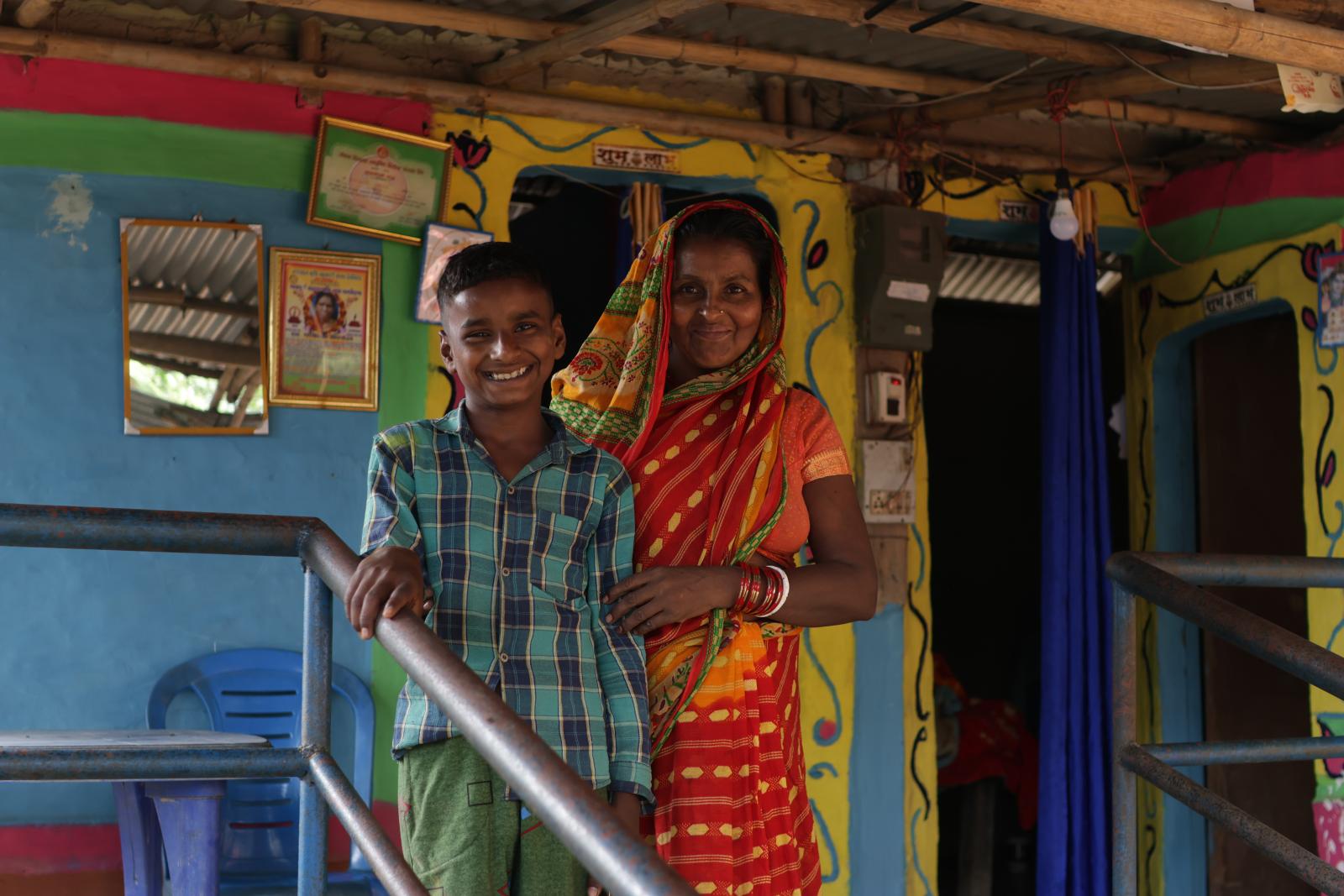 A woman in an orange and red sari stands with a boy in a blue and green plaid shirt. They are both smiling. Behind them is a colorful wall painted in blue, green, yellow, and red.