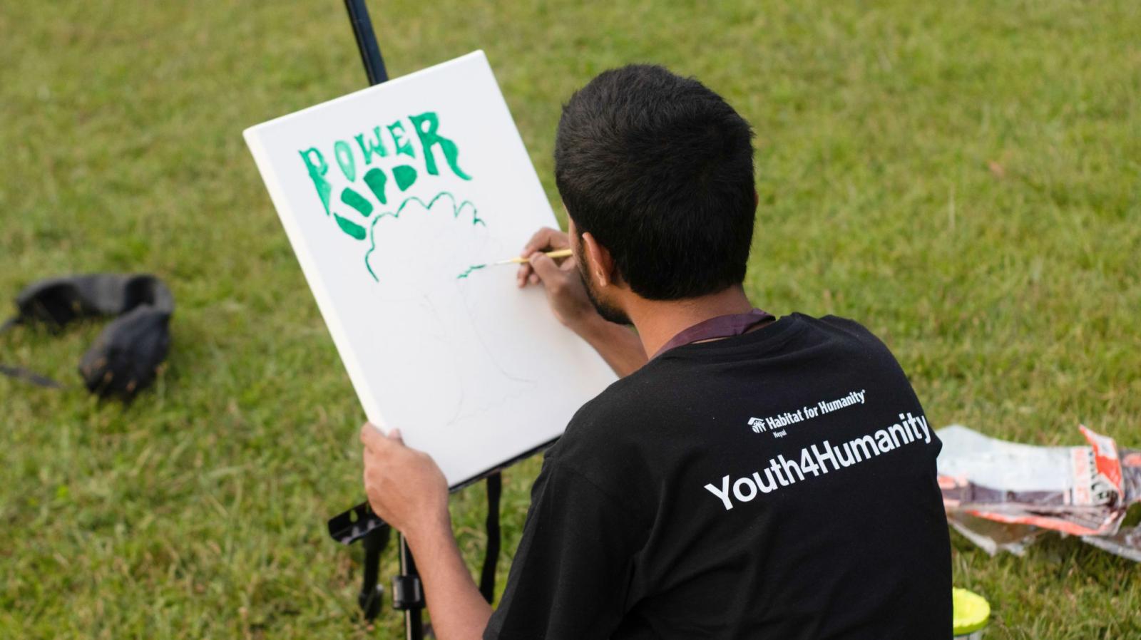A young man sits outside on the grass, painting a sign on an easel. The sign says "Power" over a drawing of a tree. The man's shirt reads "Habitat for Humanity" and "Youth4Humanity" on the back. 