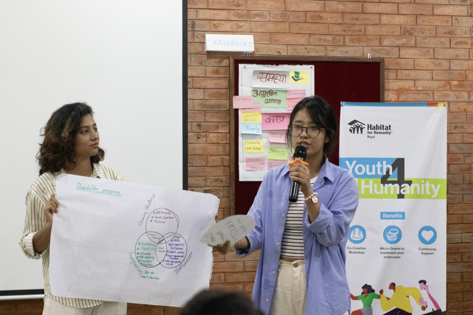 Two young women giving a presentation. One holds a poster while the other speaks into a microphone. Behind them is a Youth 4 Humanity banner with the Habitat logo. 