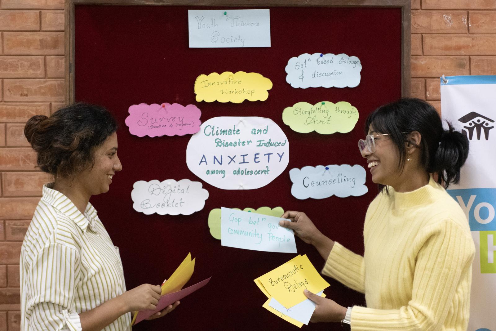 Two women smiling in front of a bulletin board with various ideas in bubbles.