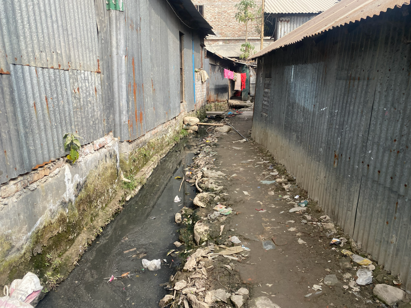 A polluted drainage ditch runs alongside a litter-strewn dirt path between rusty metal buildings in Bhashantek. Clothes hang on a line to dry further down the path.