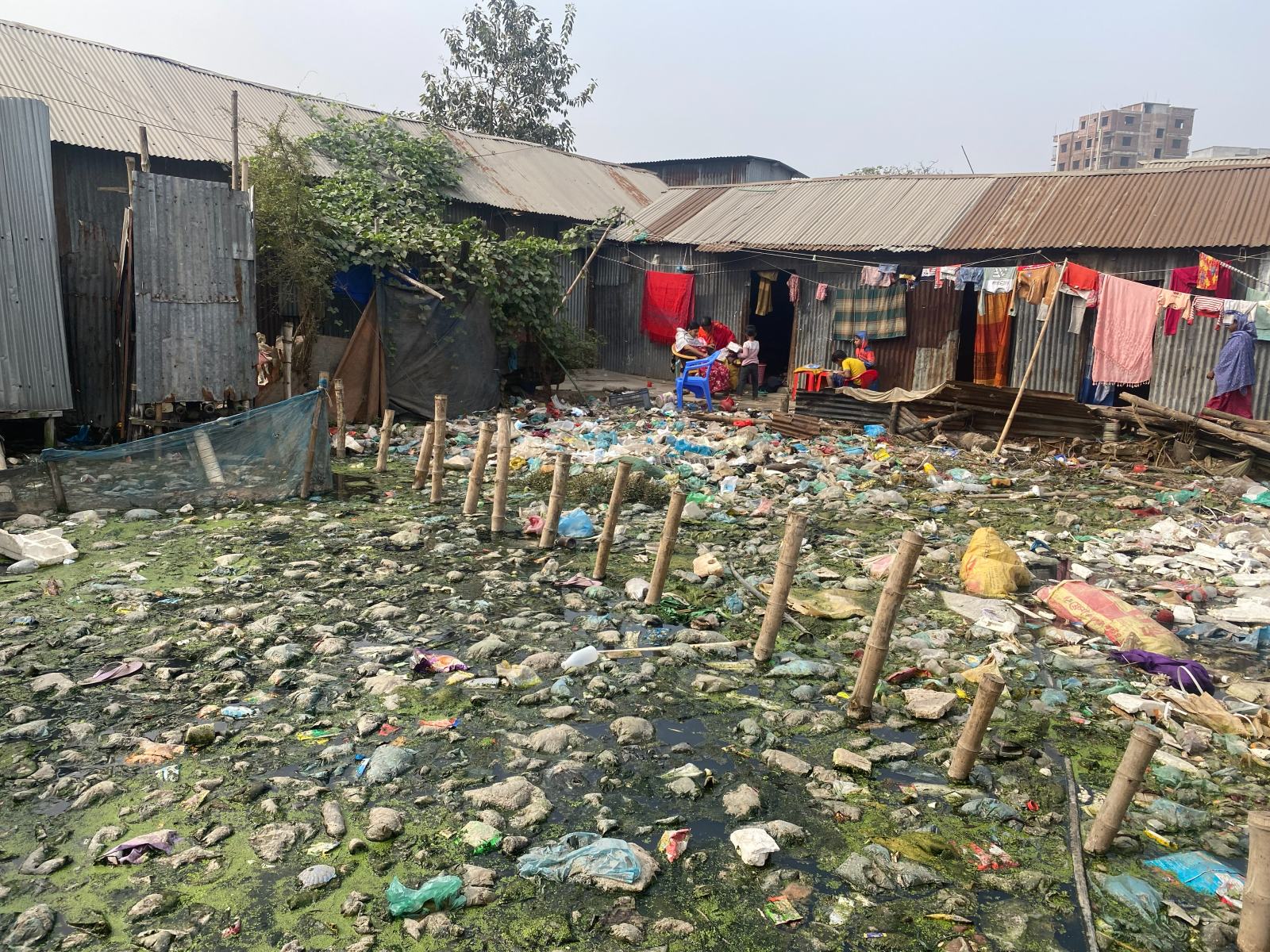 In the foreground, a swampy yard full of garbage is dissected by a bamboo fence. In the background, women and children gather on a narrow concrete strip outside their home, beneath lines of drying laundry. The buildings are made from a patchwork of corrugated metal.