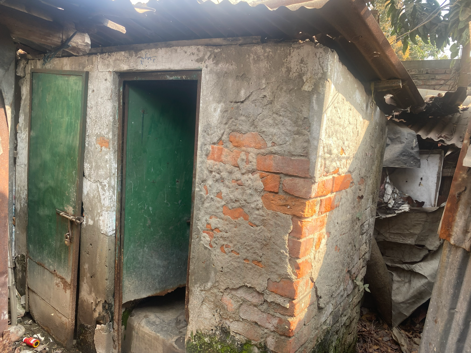 A ramshackle brick and concrete latrine building with two green doors and a corrugated metal roof. 