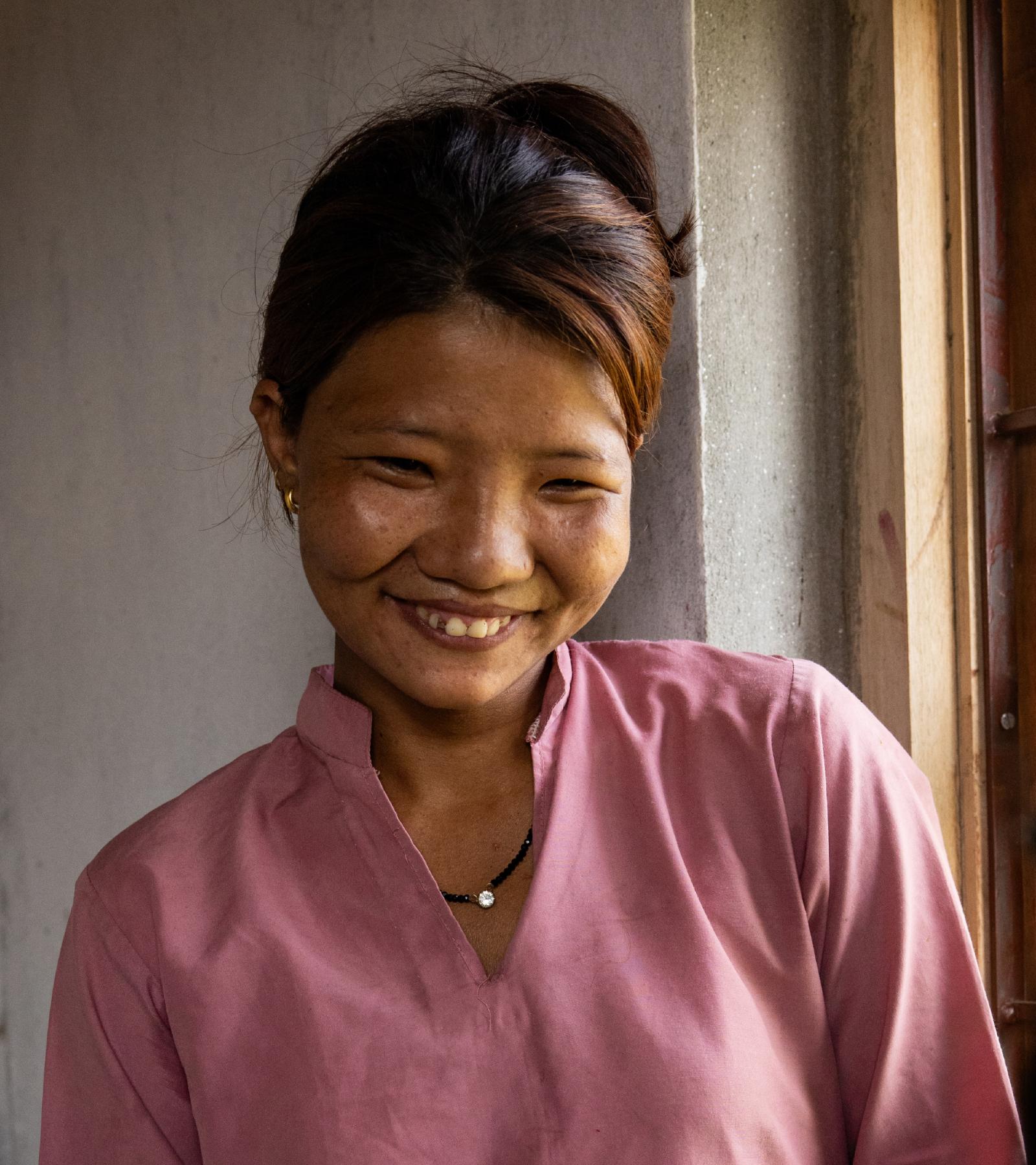 Kusum smiles and looks down. She is wearing a pink button-down long-sleeved shirt.
