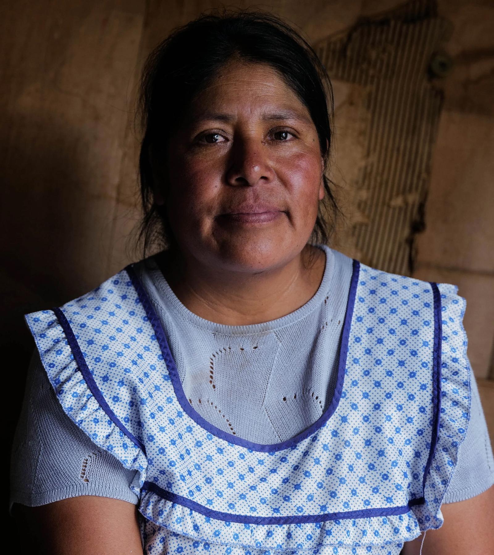 A woman sits in a blue apron with a solemn expression on her face.