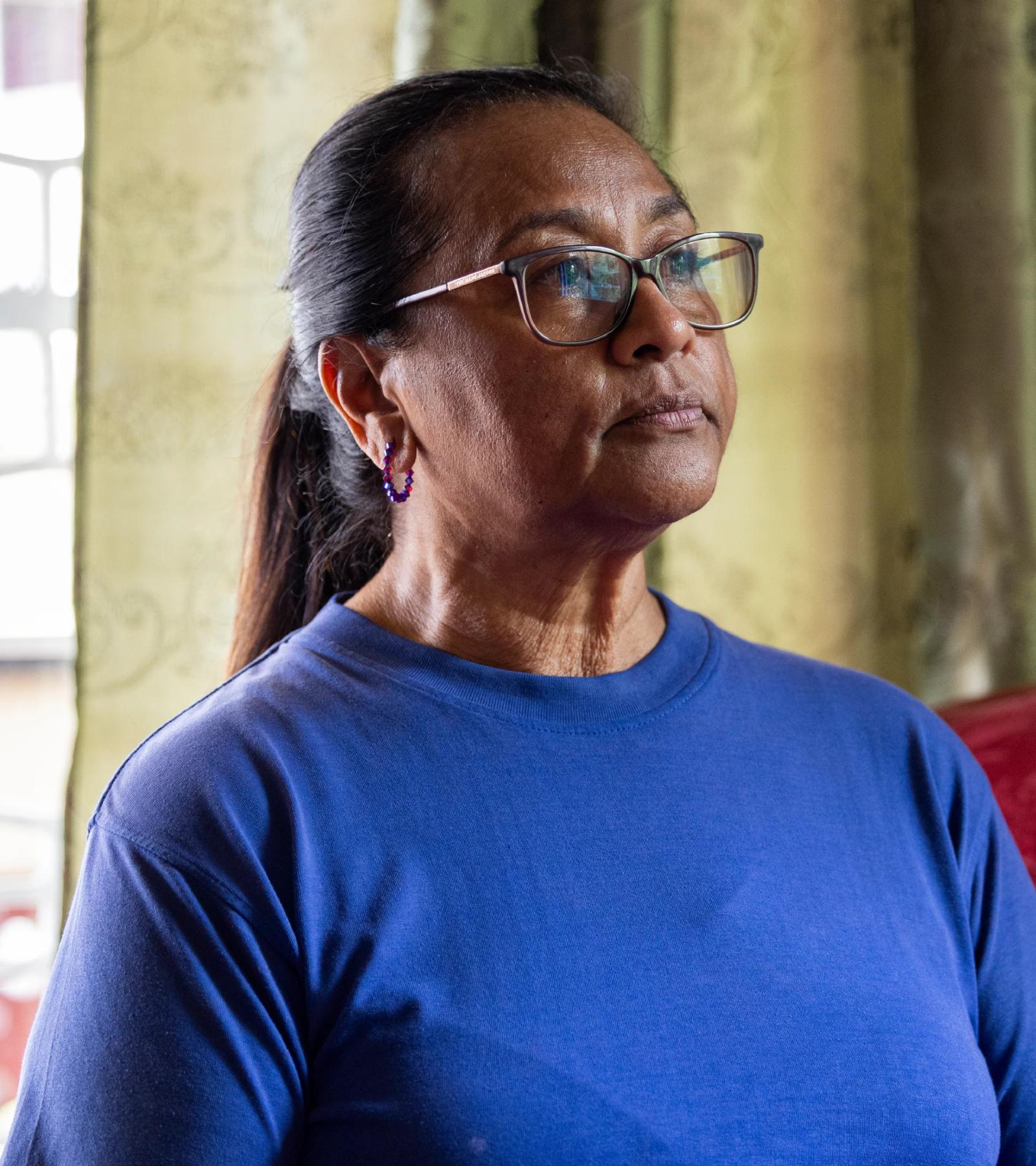 A woman in a blue shirt and glasses has a somber expression on her face as she looks off camera.