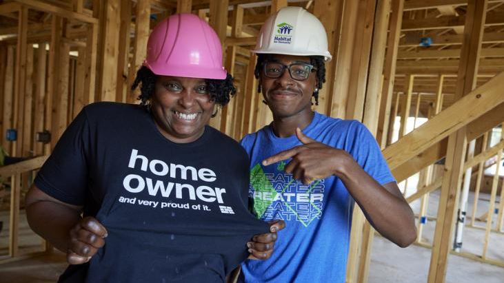 A Black woman in a hardhat smiles inside an unfinished Habitat house in progress, showing her "Homeowner and proud of it" shirt, while a young Black man in a hardhat points to it.